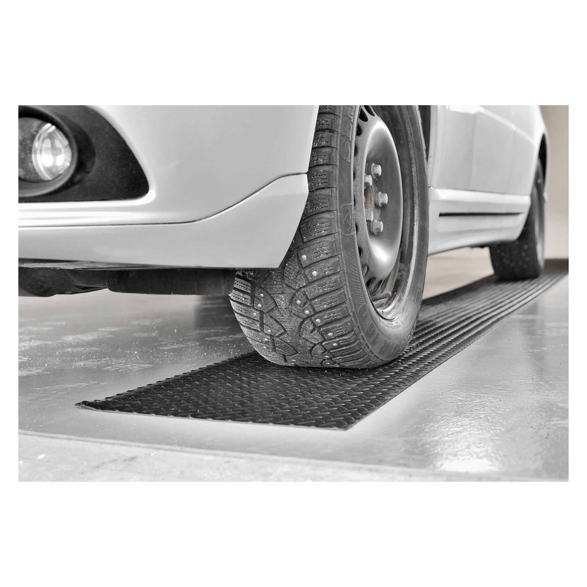 En närbild av ett bildäck med vintermönster som vilar på en Hamron Floor Protection-matta i ett garage, med framstötfångaren och en del av hjulhuset också i bild.