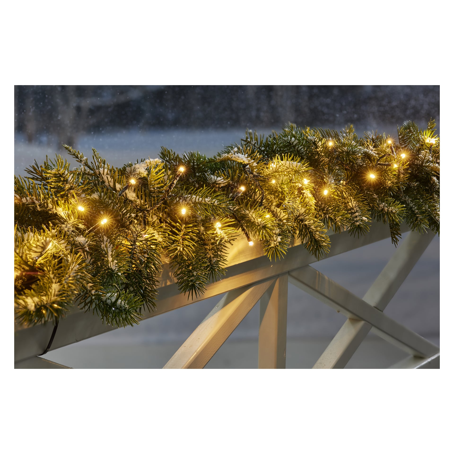 En EKVIP tallgirland dekorerad med LED-ljusslingor är lindad runt ett vitt räcke, med snö som lagt sig på grönskan och mer snö synlig bakom.