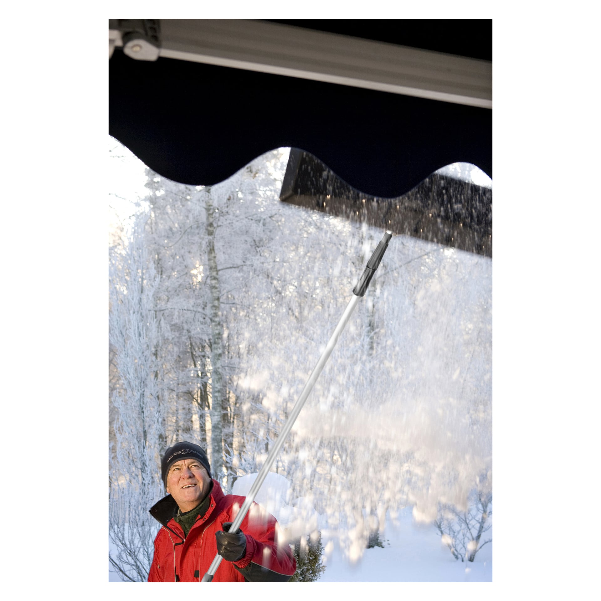 En person i röd jacka och svart hatt använder Hard Head Telescopic Roof Rake för att ta bort snö från ett tak en snöig vinterdag, med snötäckta träd synliga bakom sig.
