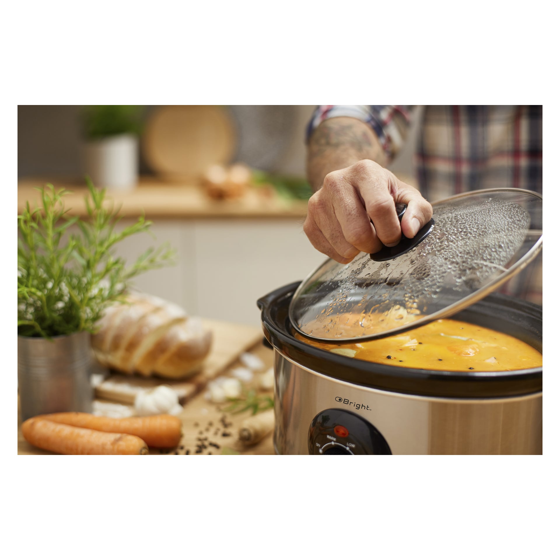 Någon lyfter locket från en Bright-slow cooker med apelsinsoppa. På köksbänken i trä finns morötter, en limpa bröd, vitlök och en krukväxt, allt arrangerat för att antyda förberedelser.