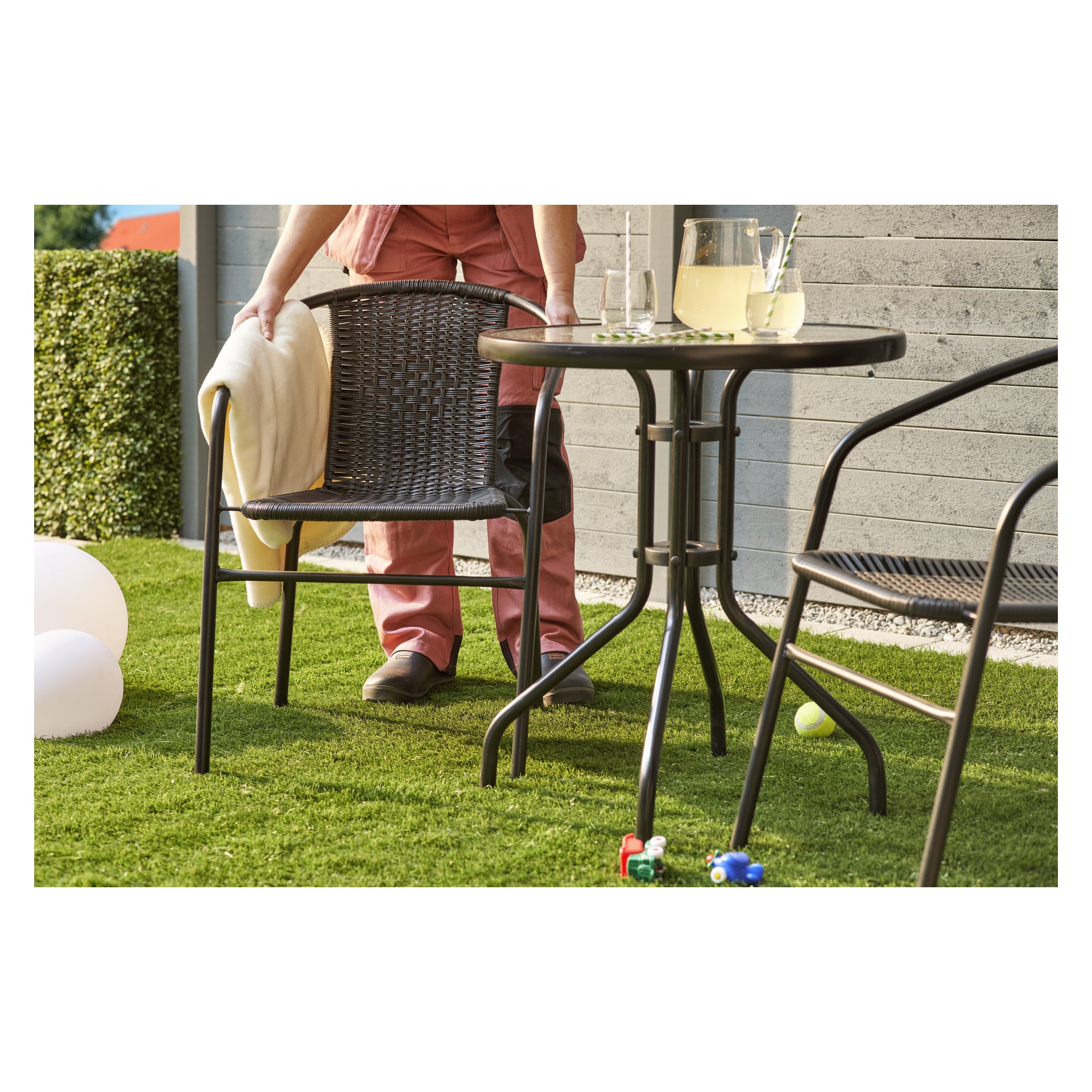 En person i rosa legger et lett teppe over en svart vevd EKVIP Café-stol ved siden av et rundt bord med limonade og glass. Scenen foregår på grønt gress, med leker og ballonger synlige i nærheten.