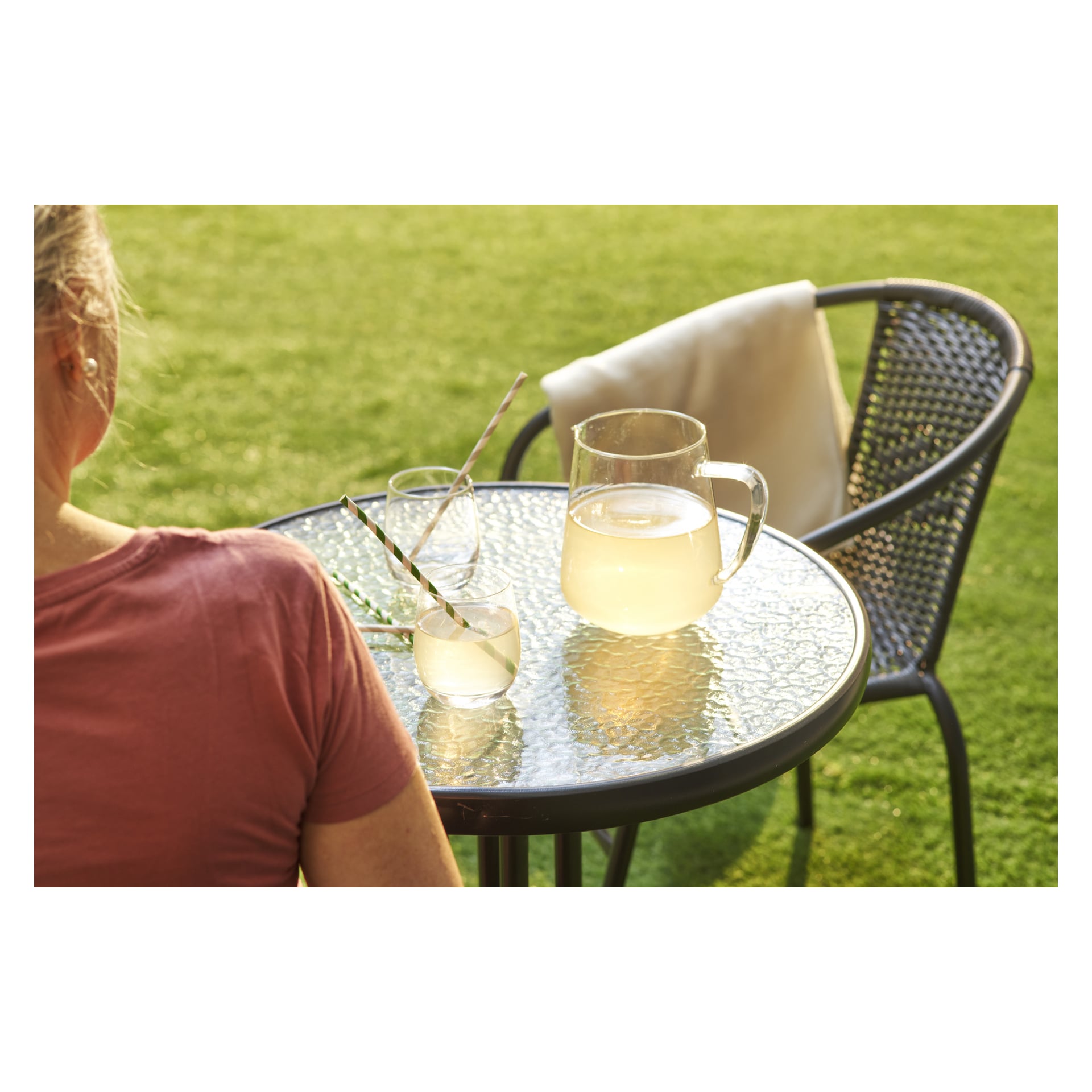 En person slapper av ved et glassbord ute dekket med en mugge og to glass limonade, begge med stripete sugerør. En EKVIP Café-stol står like ved på den grønne plenen, sollyset strømmer over den og en lett klut hviler over ryggen.