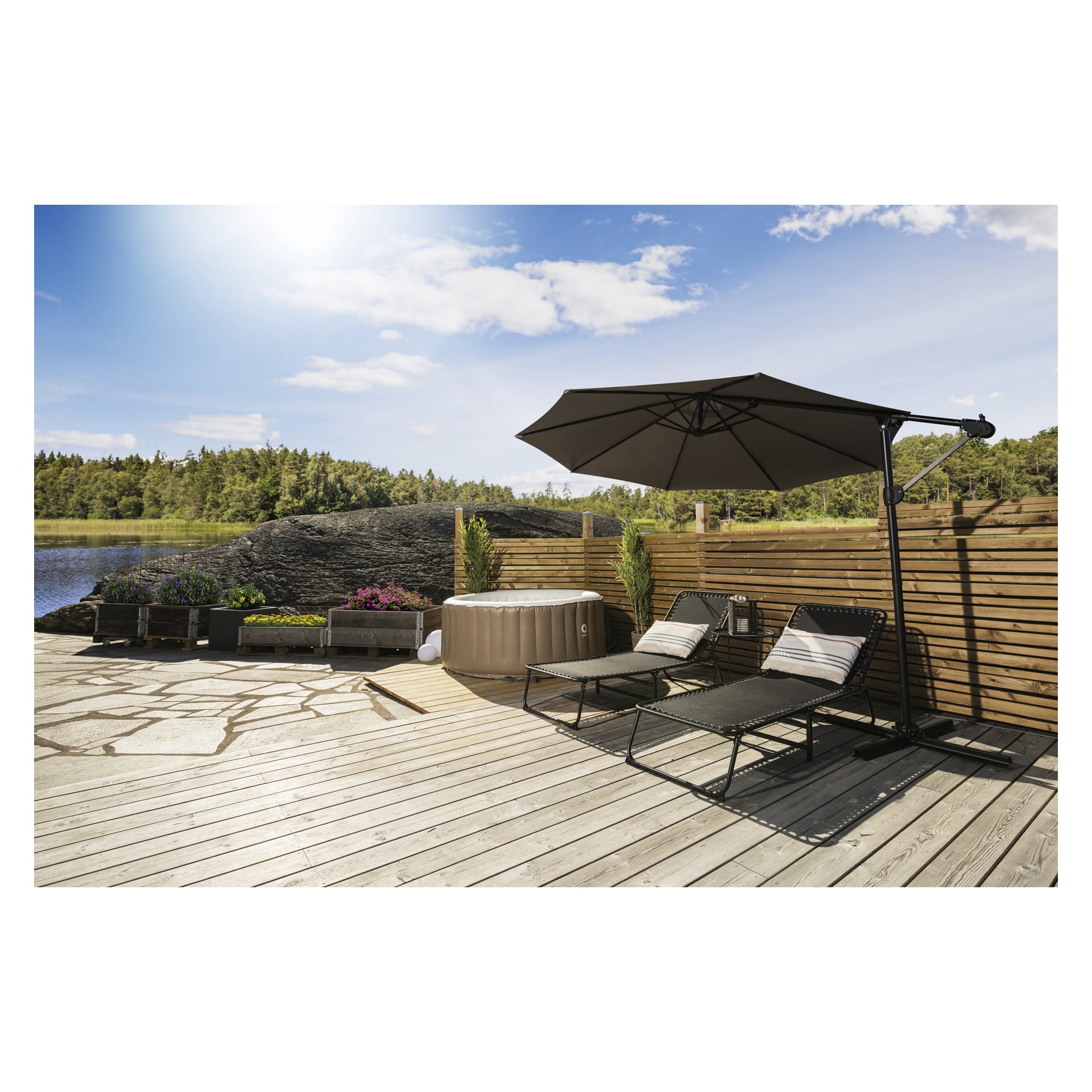 På en träaltan står två svarta vadderade solstolar bredvid en rund bubbelpool och planteringslådor fyllda med blommor, inramade av ett trästaket för avskildhet. Ett stort EKVIP-parasoll erbjuder skugga, och i bakgrunden finns en sjö med träd under en klarblå himmel.