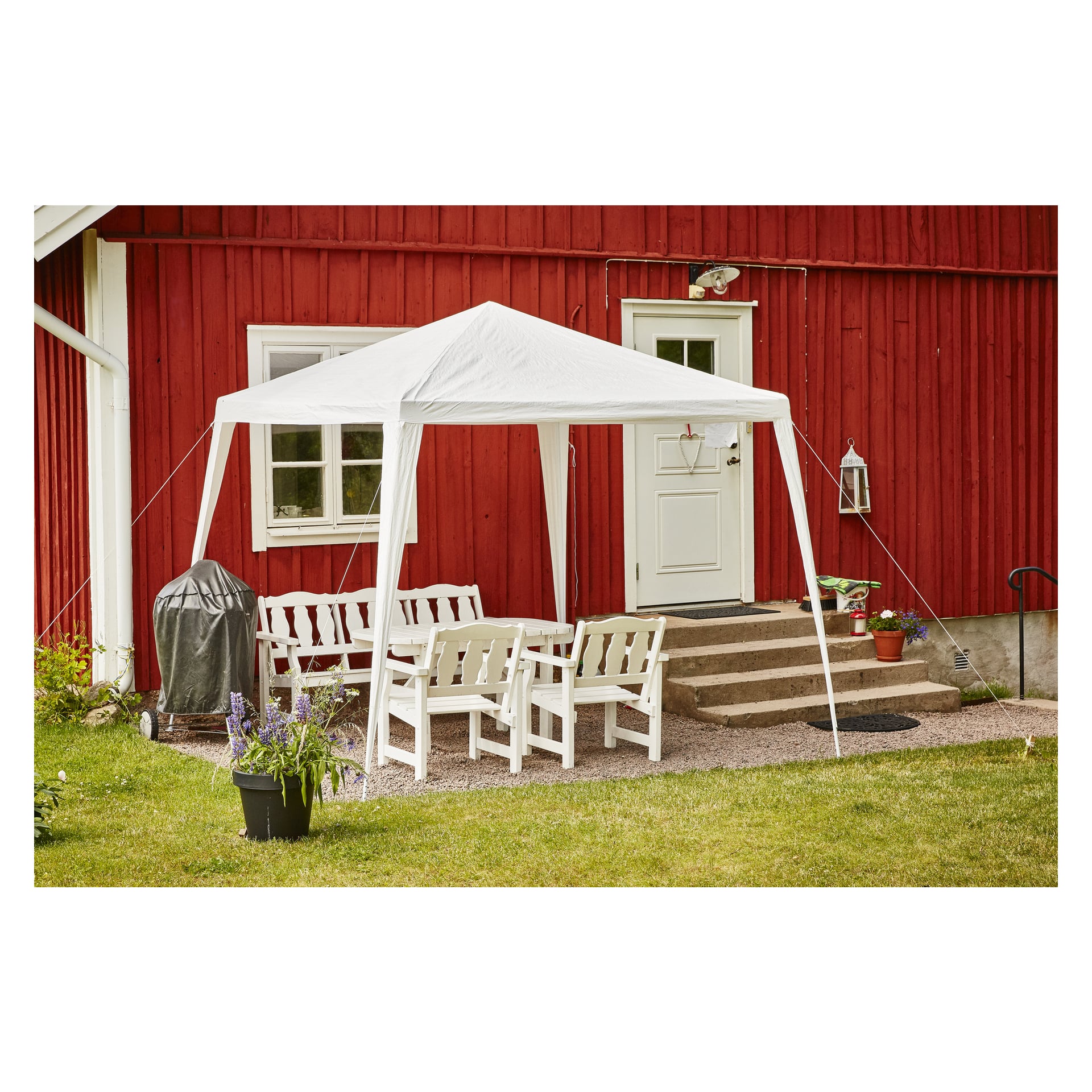 Ett EKVIP Party tält skuggar en samling vita utomhusstolar och ett bord på en grusterrass bredvid ett rött trähus, med trappor, dörr, flera krukväxter och en täckt grill. I förgrunden finns gräs och en svart kruka.
