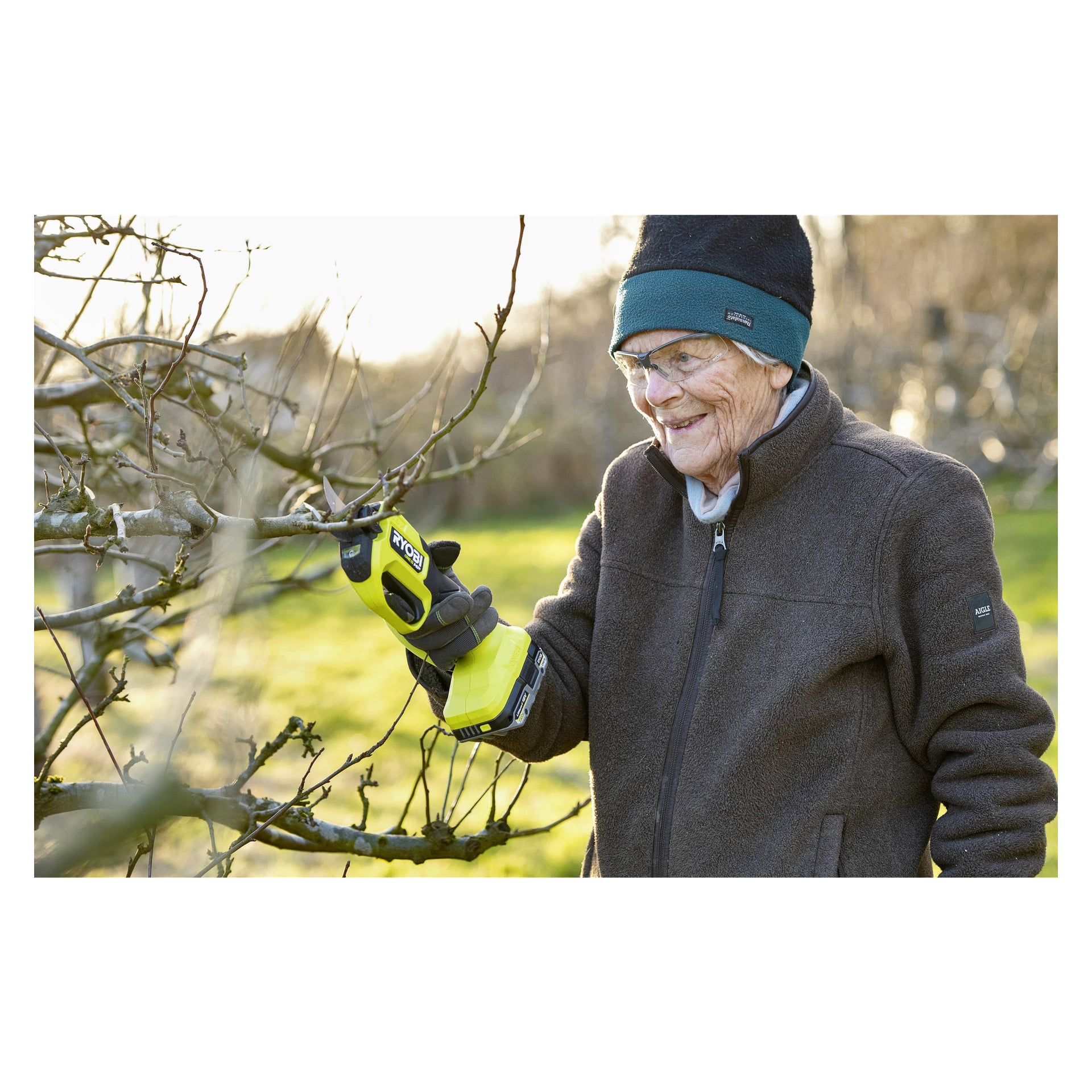 En äldre person klädd i jacka, handskar, hatt och skyddsglasögon är utomhus en solig dag och använder Ryobi One+ HP batteridrivna sekatörer RY18SCXA-0 för att trimma trädgrenar.