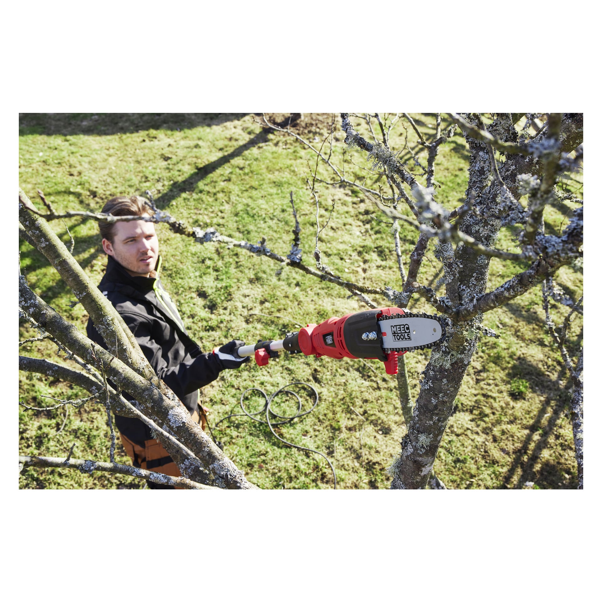 En man i handskar och svart jacka använder en röd elektrisk beskärningssåg från Meec Tools för att såga lavtäckta grenar från ett träd i en gräsbevuxen utomhusmiljö.