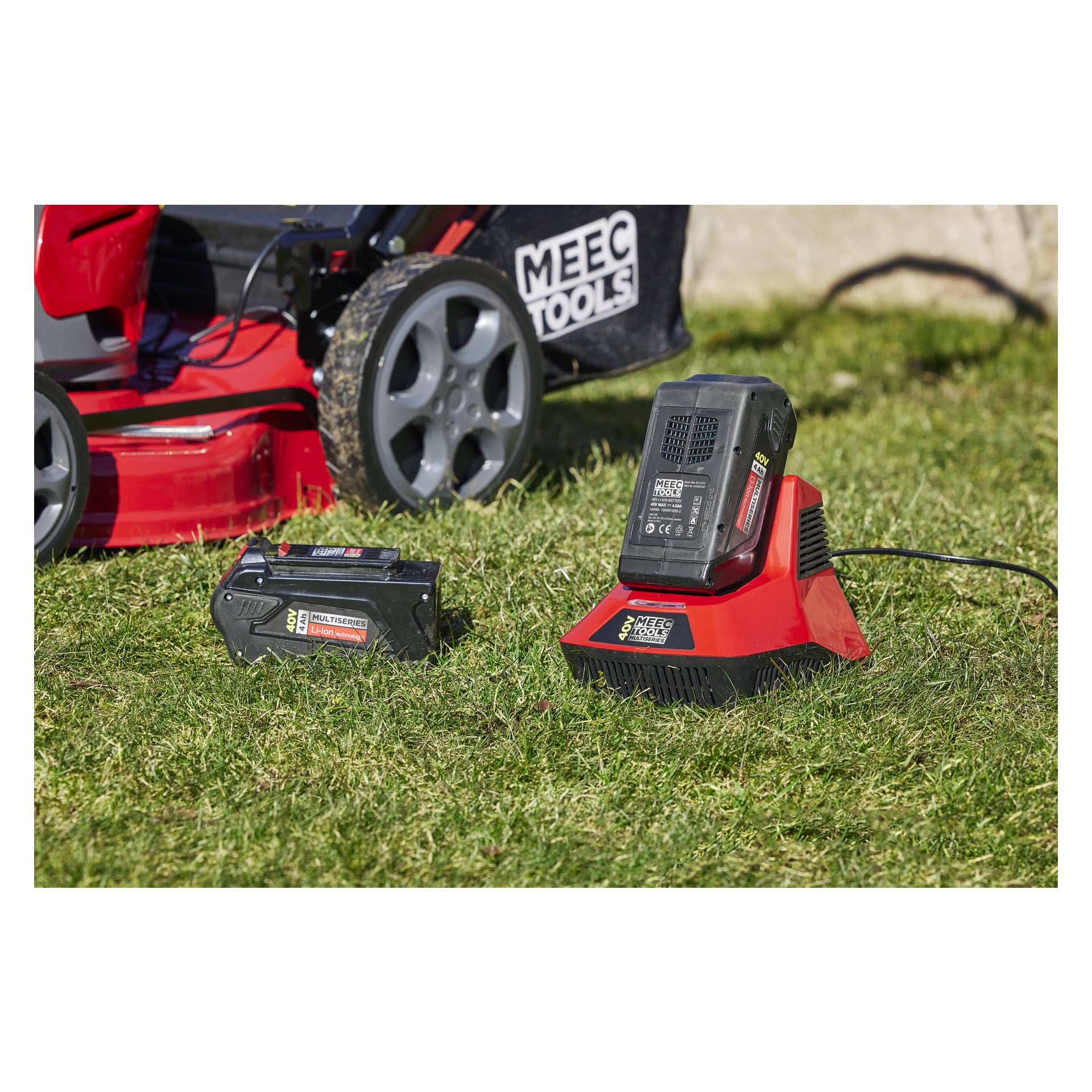Ett svart batteri och en röd Meec Tools Multiseries 40 V snabbladdare vilar på grönt gräs i starkt solljus, med en röd sladdlös gräsklippare synlig i bakgrunden.