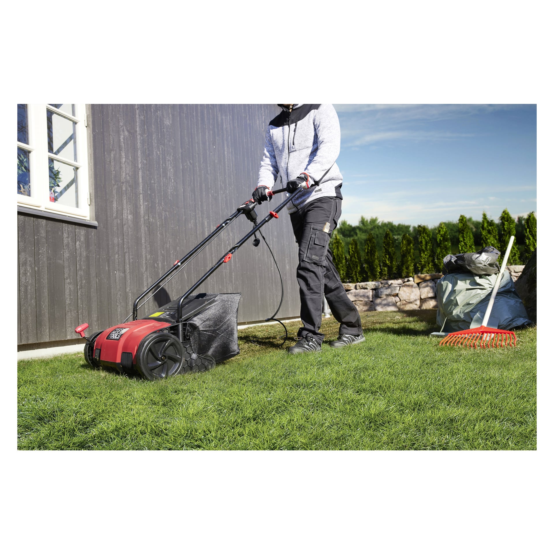 En person klädd i en grå hoodie och svarta byxor använder Meec Tools Scarifier 2-i-1 på en gräsmatta bredvid ett hus. I närheten finns en kratta och en påse fylld med trädgårdsskräp.