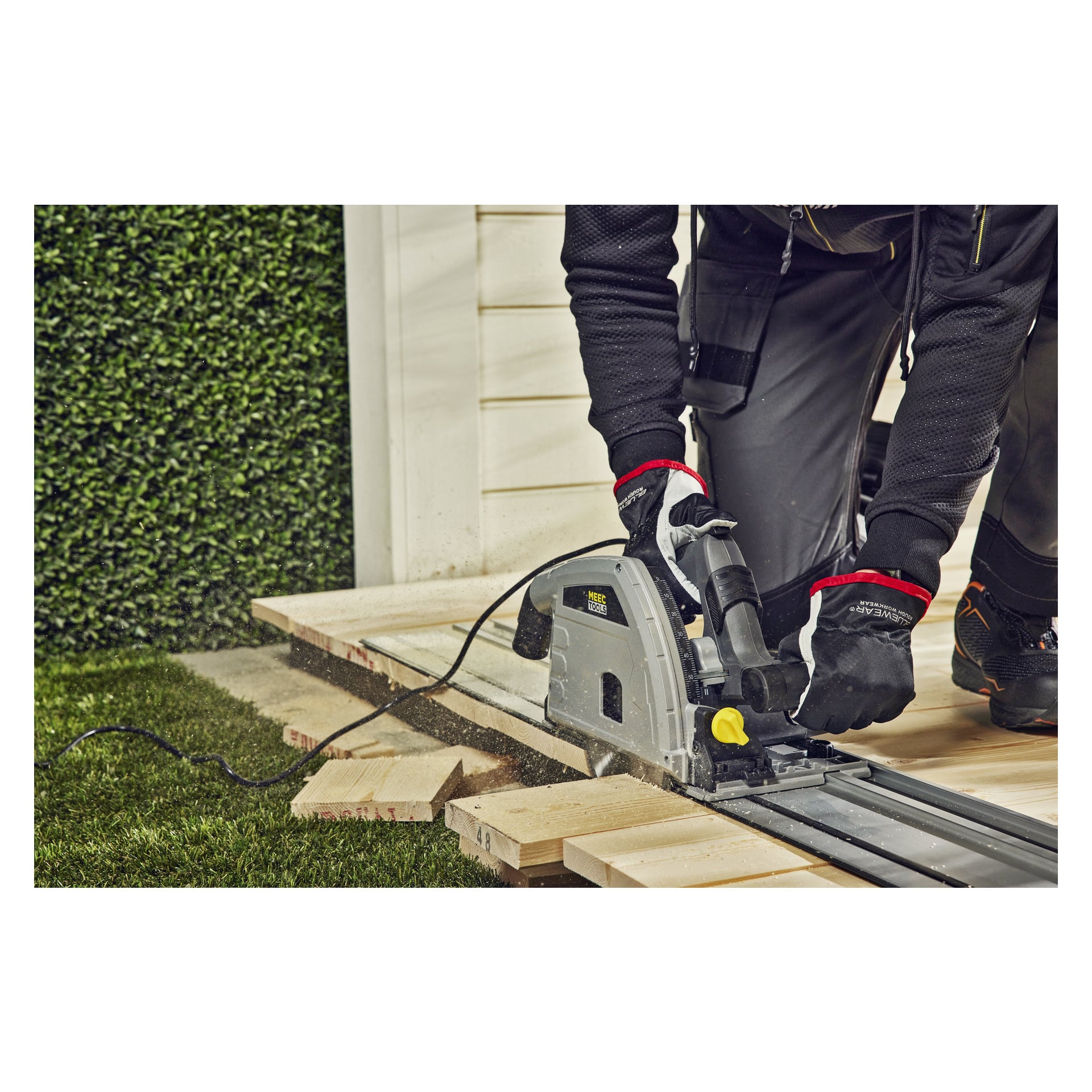 Handskbeklädda händer manövrerar en Meec Tools Plunge-såg CI-W HOME 300 med styrskena, där träplankor sågas utomhus via gräs och en vit konstruktion.