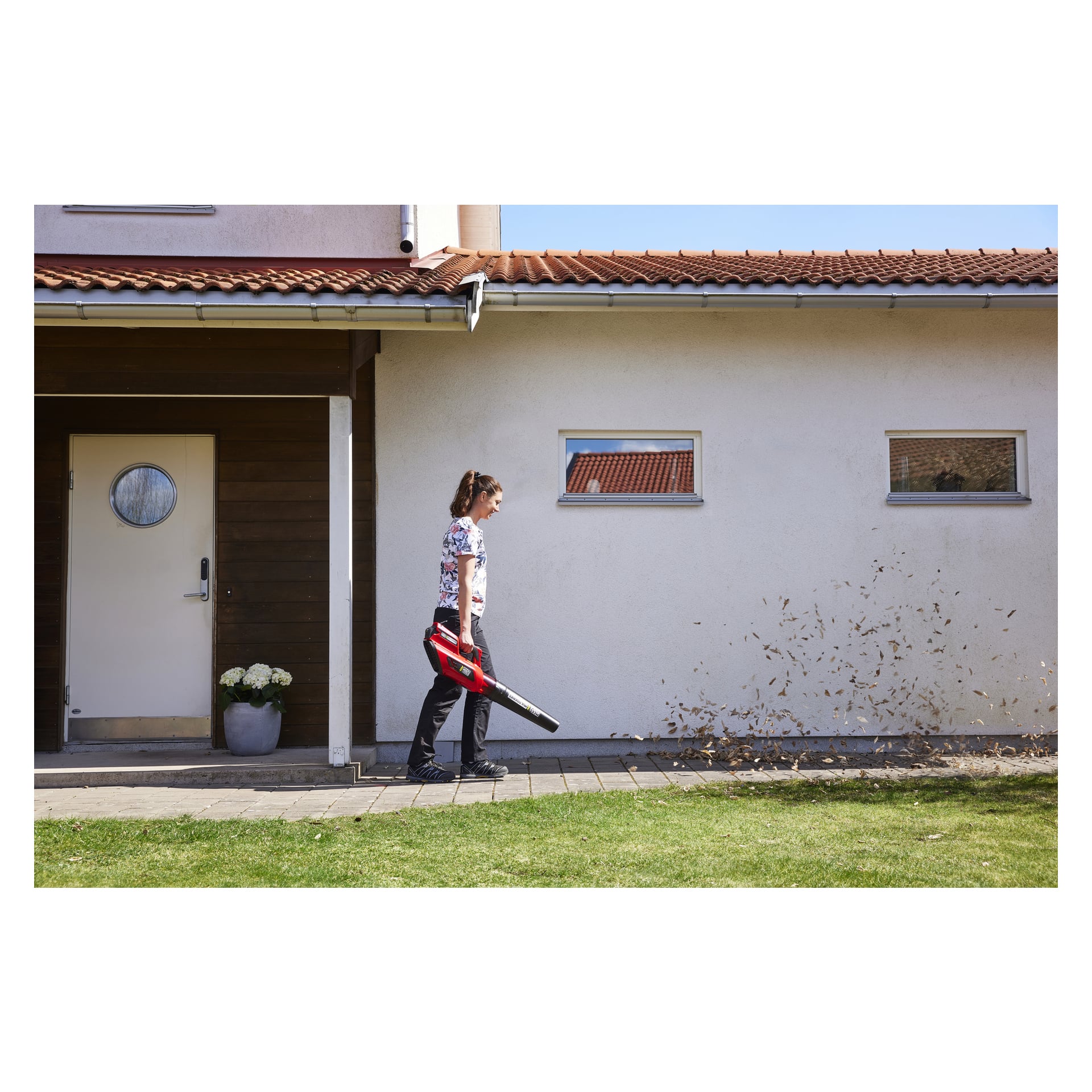 En person kör en röd och svart Meec Tools Multiseries 40 V batteridriven lövblåsare på en asfalterad gångväg utanför ett hus med både vita och träutvändiga sektioner, och blåser löv åt höger. Det finns en krukväxt nära dörröppningen och två små fönster på väggen.