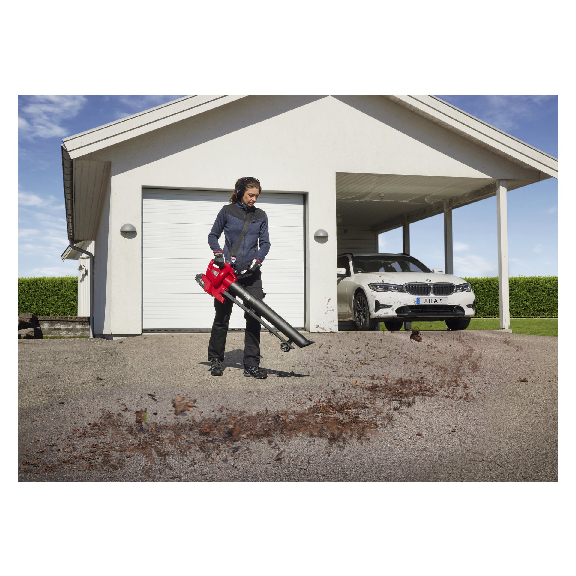 En person kör en Meec Tools Multiseries 40 V lövblås/dammsugare på en uppfart framför ett vitt hus med garage, där löv och skräp röjs medan en vit bil står under carporten.