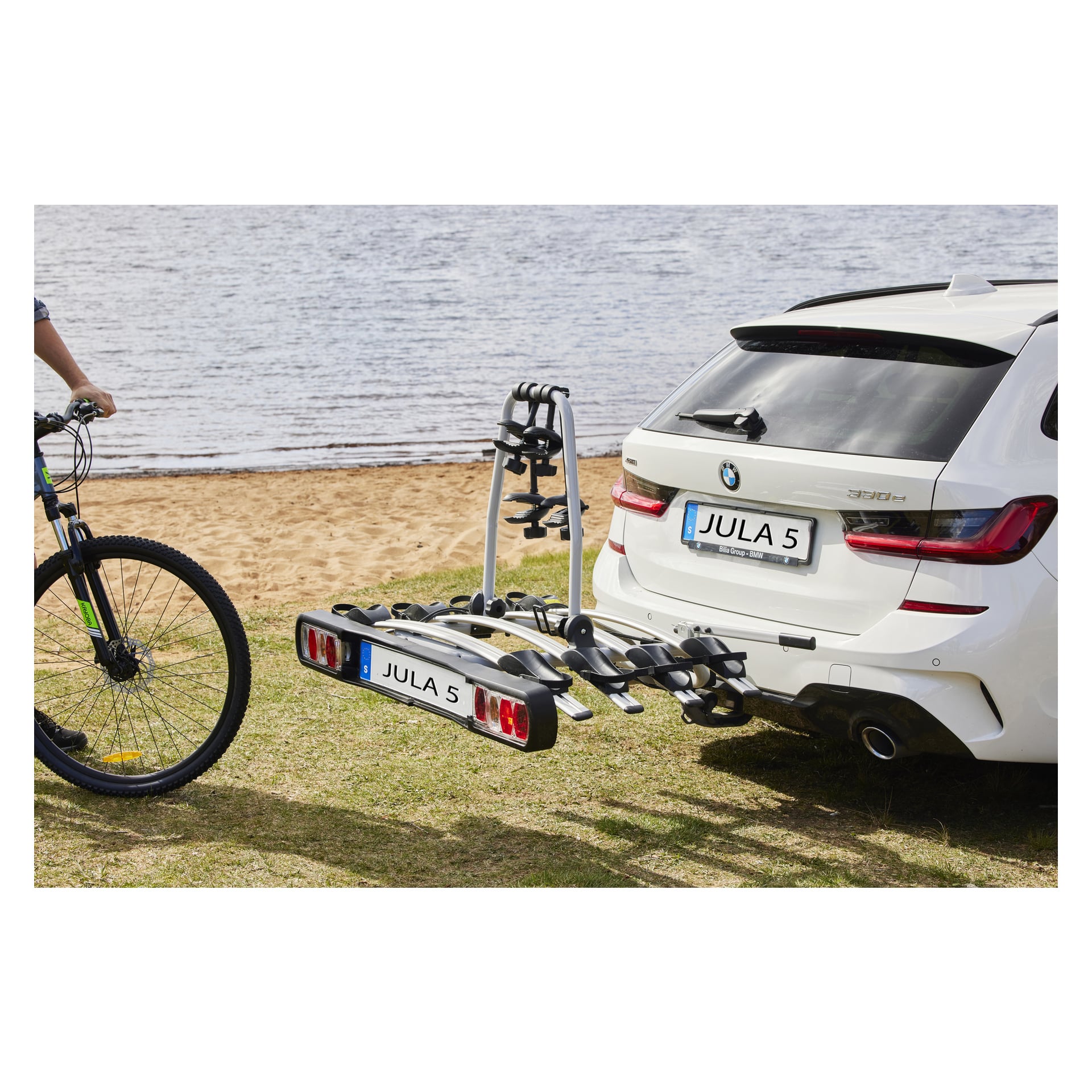 Białe BMW 330e kombi stoi przy nabrzeżu, a z tyłu montuje się bagażnik na drążku holowniczym Hamron. Obok pojazdu stoi osoba trzymająca rower.
