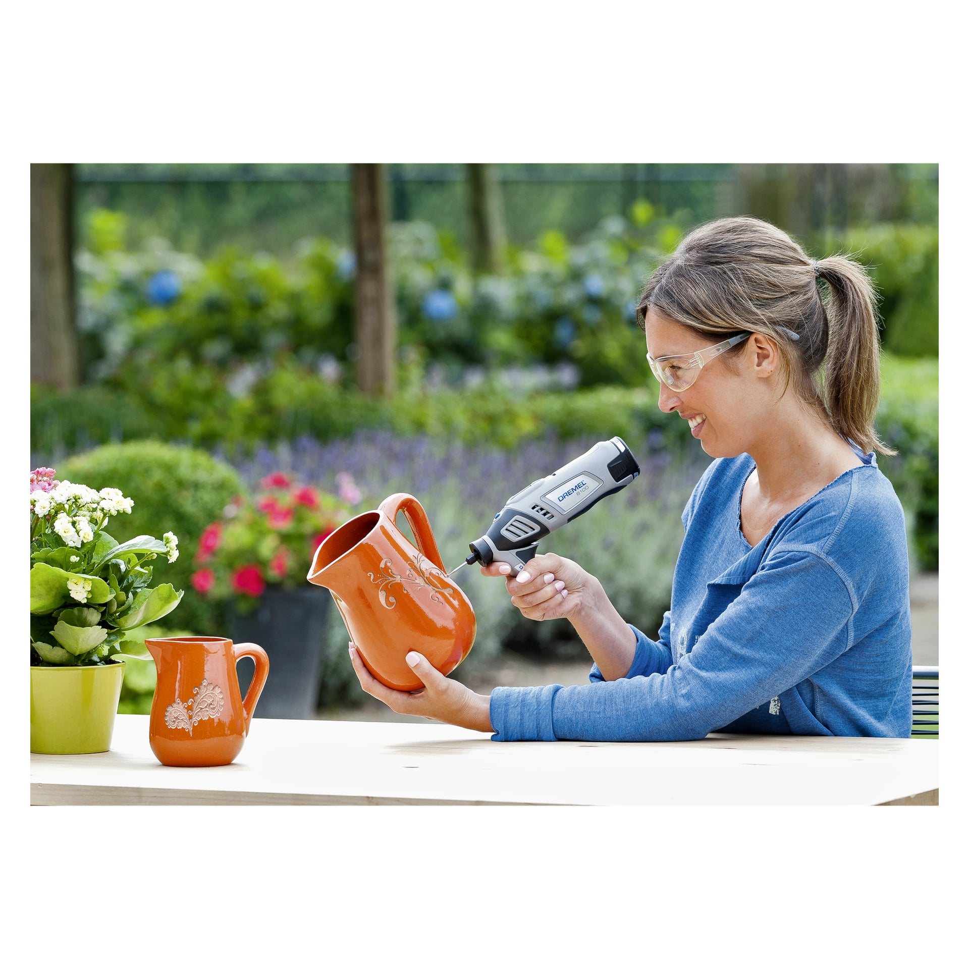 En kvinne med vernebriller bruker et Dremel Precision-håndtak 577 roterende verktøy for å gravere blomstermønstre på en oransje keramikkmugge ved et utebord, omgitt av blomster og en mindre, matchende mugge.
