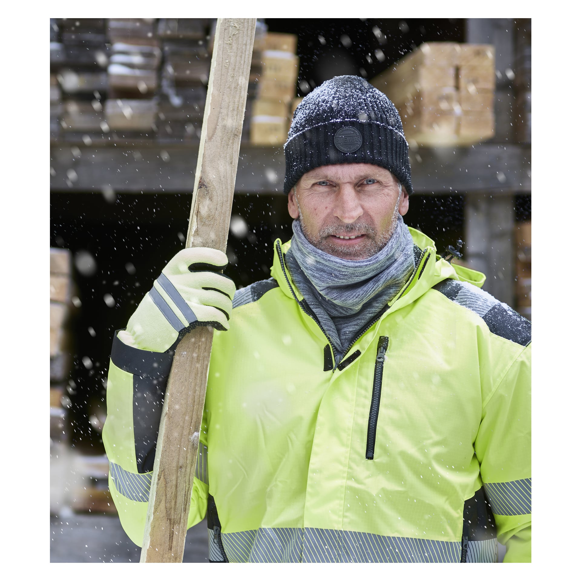 Ein Mann, gekleidet in einen Kayoba Outdoor-Halskragen, Handschuhe und eine schwarze Wintermütze, trägt ein Holzbrett auf der Schulter, während um ihn herum Schnee fällt. Im Hintergrund sind Holzstaple zu sehen.