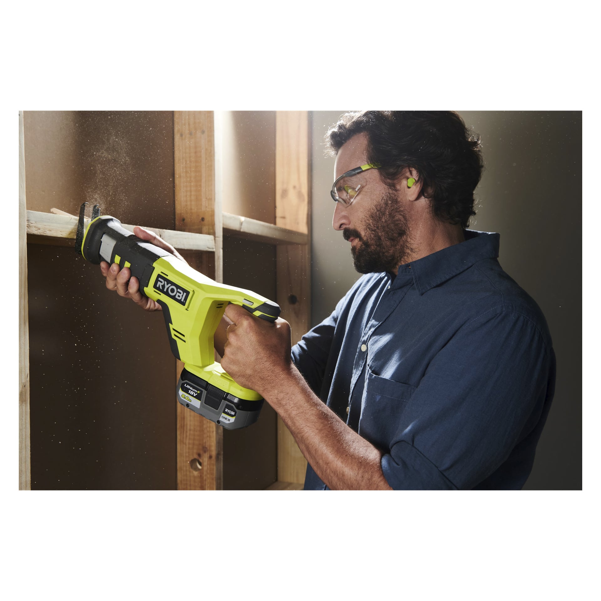 En man, utrustad med skyddsglasögon och öronproppar, använder en Ryobi One+ sladdlös bajsåg (modell RRS18-0) för att skära igenom en träskiva fäst vid en ofärdig vägg. Sågspån kan ses sväva i luften.