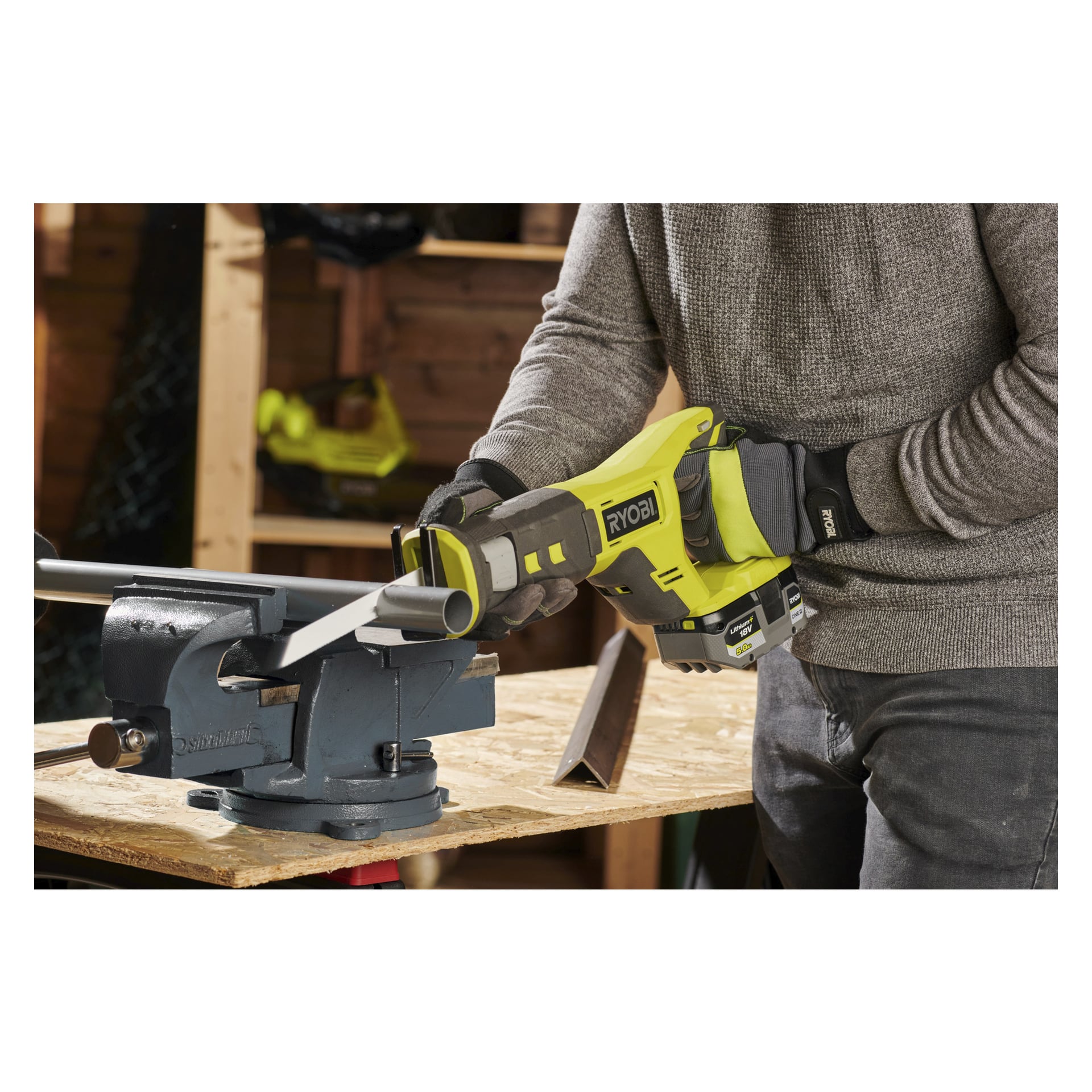 Handskbeklädda händer manövrerar en gul och svart Ryobi One+ batteridriven bajonsåg (RRS18-0) för att såga igenom ett metallrör som klämts fast i ett blått skruvstäd på en arbetsbänk inne i en verkstad.