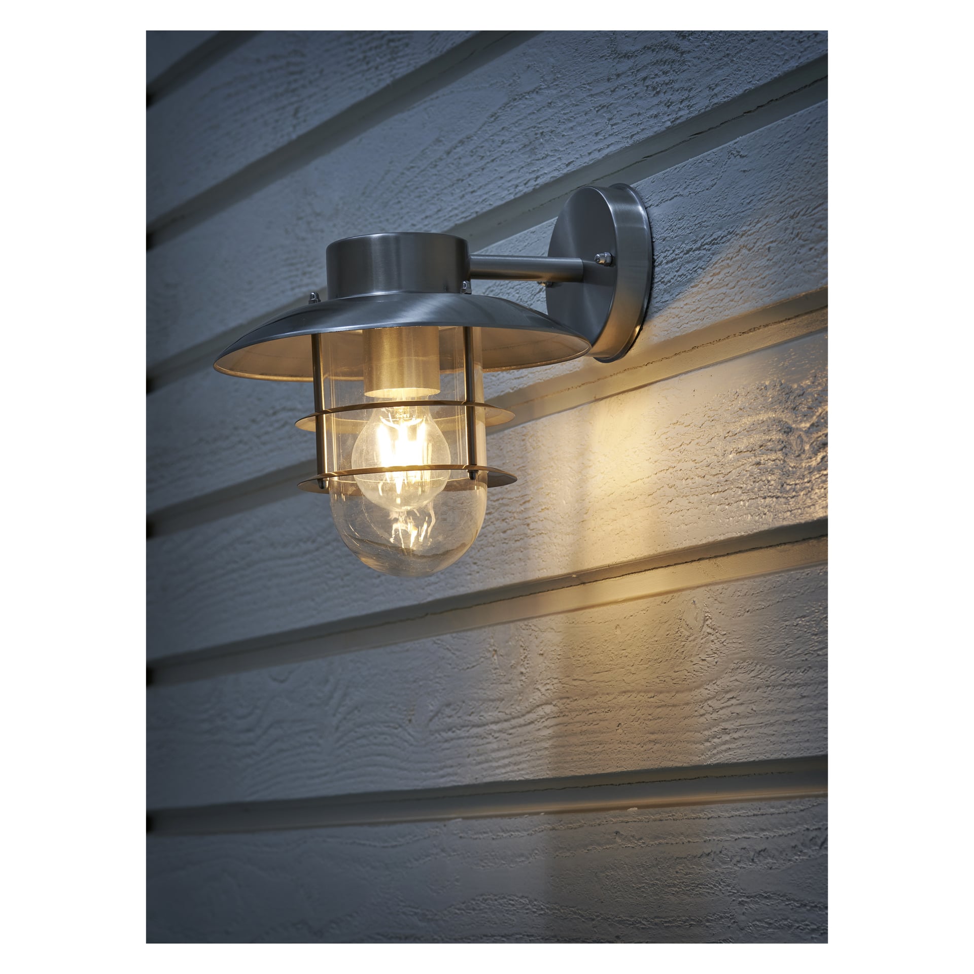 En EKVAP-vägglampa med metallram och exponerad klar lampa är fäst på en ljus, horisontellt panelad yttervägg och belyser den texturerade ytan med ett varmt ljus.