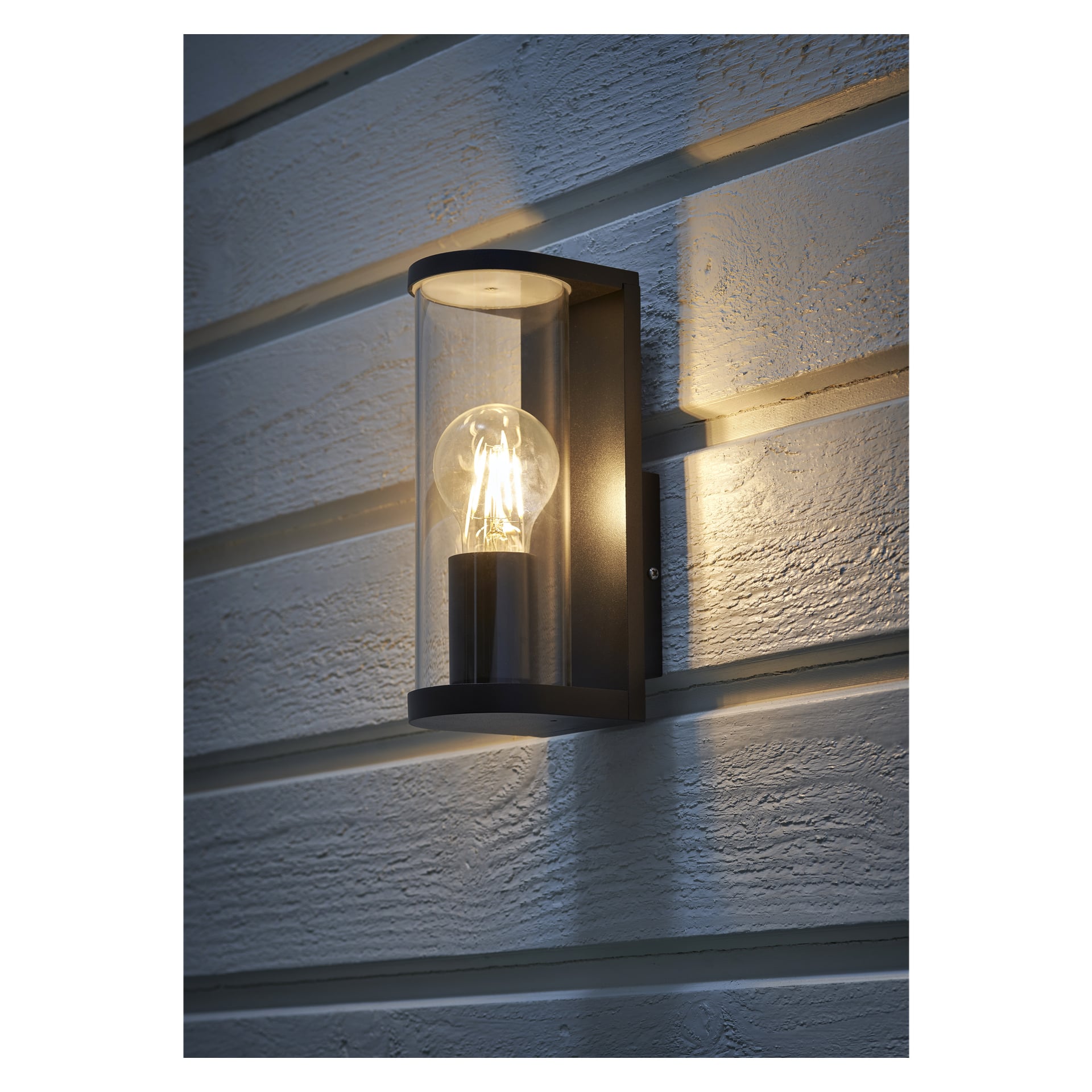 En lysande EKVIP vägglampa med ett genomskinligt cylindriskt lock är fäst på horisontell, strukturerad träpanel utomhus vid skymningen och visar den synliga lampan inuti.