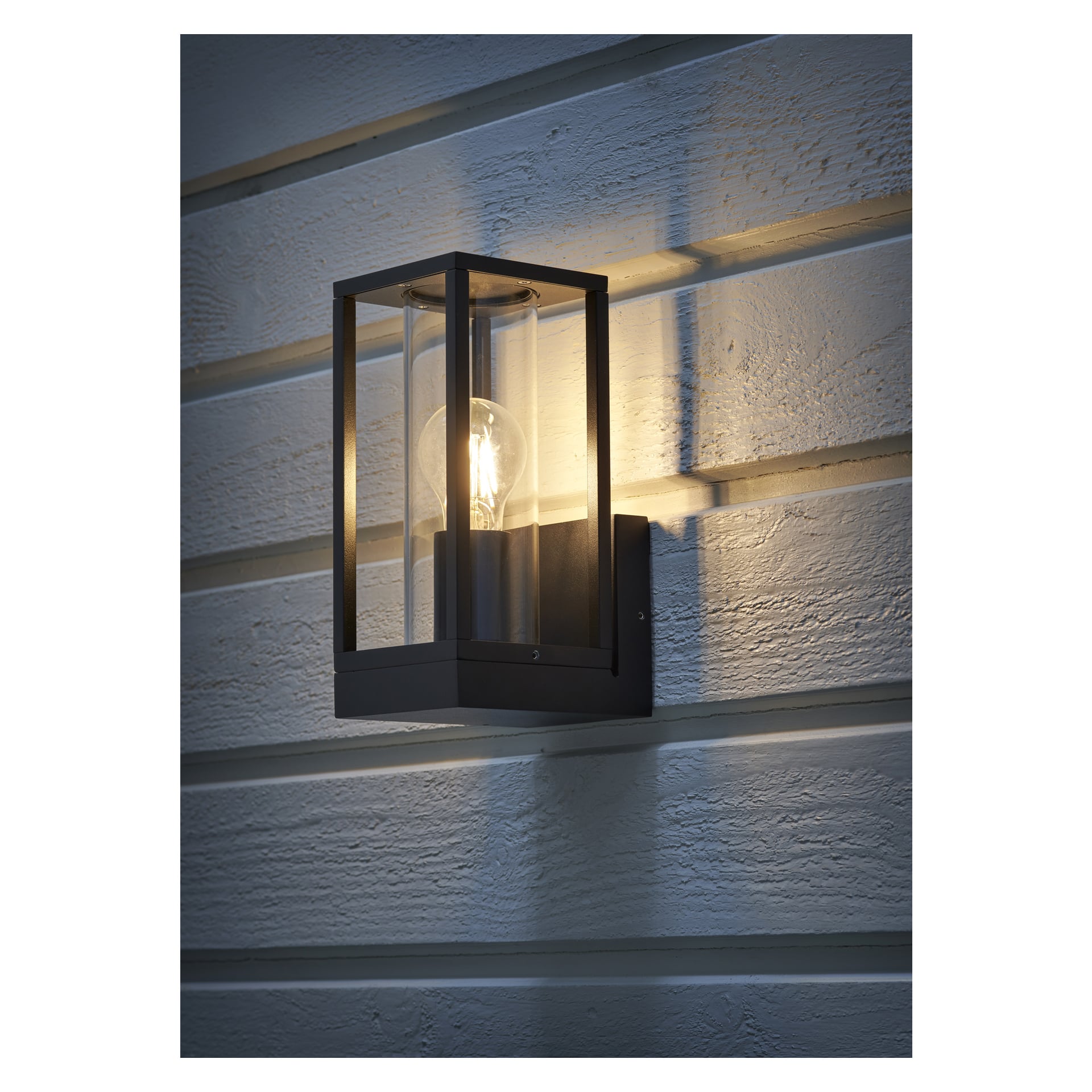 En EKVAP-vägglampa med svart metallram och klart glasskydd är fäst på en horisontell träpanelvägg och lyser upp utomhusutrymmet med ett varmt ljus.
