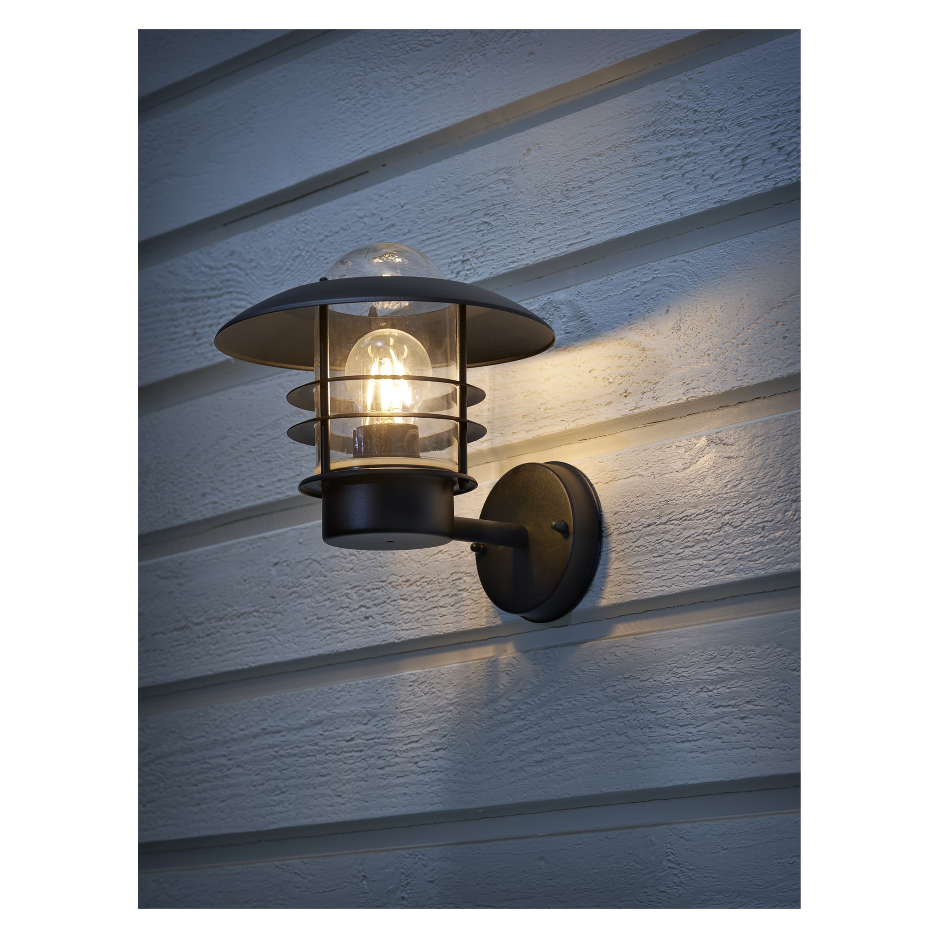 En EKVIP-vägglampa med svart metallram och glashölje är fäst på ljus horisontell fasad och lyser upp väggen med ett mjukt, varmt ljus.