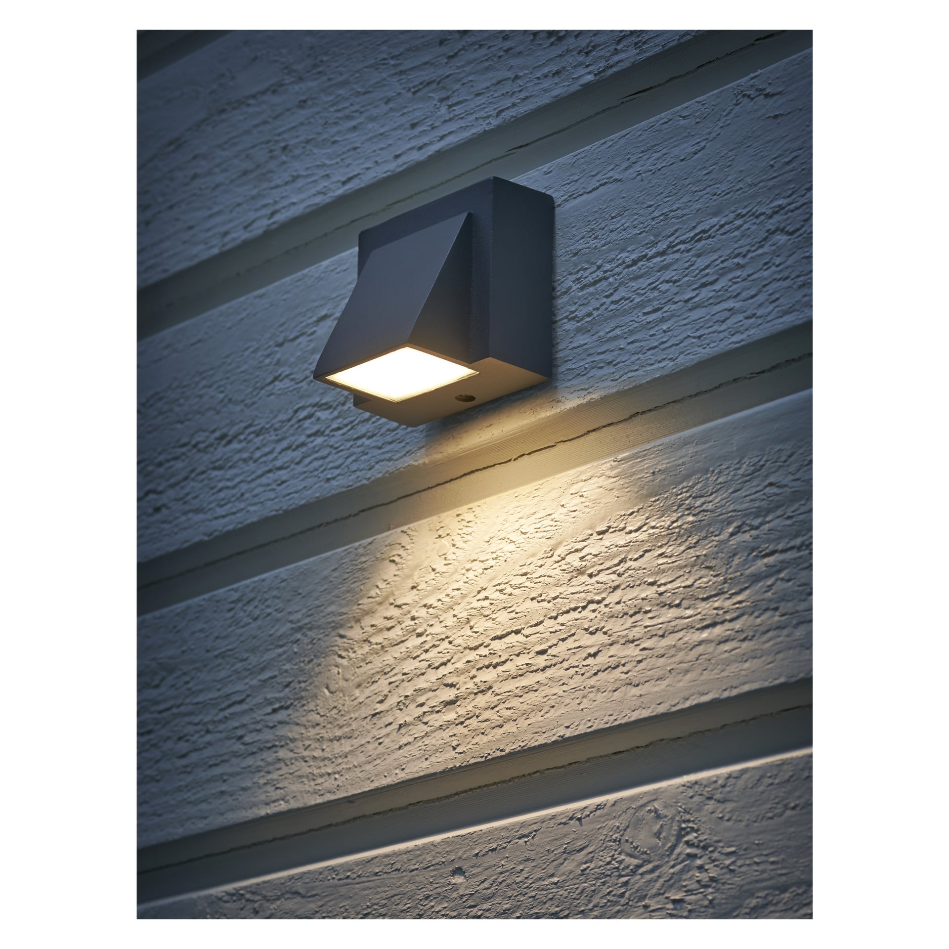 En modern, fyrkantig EKVIP Wall Spotlight LED avger ett varmt ljus som framhäver texturen av horisontell fasad under skymning eller natt.
