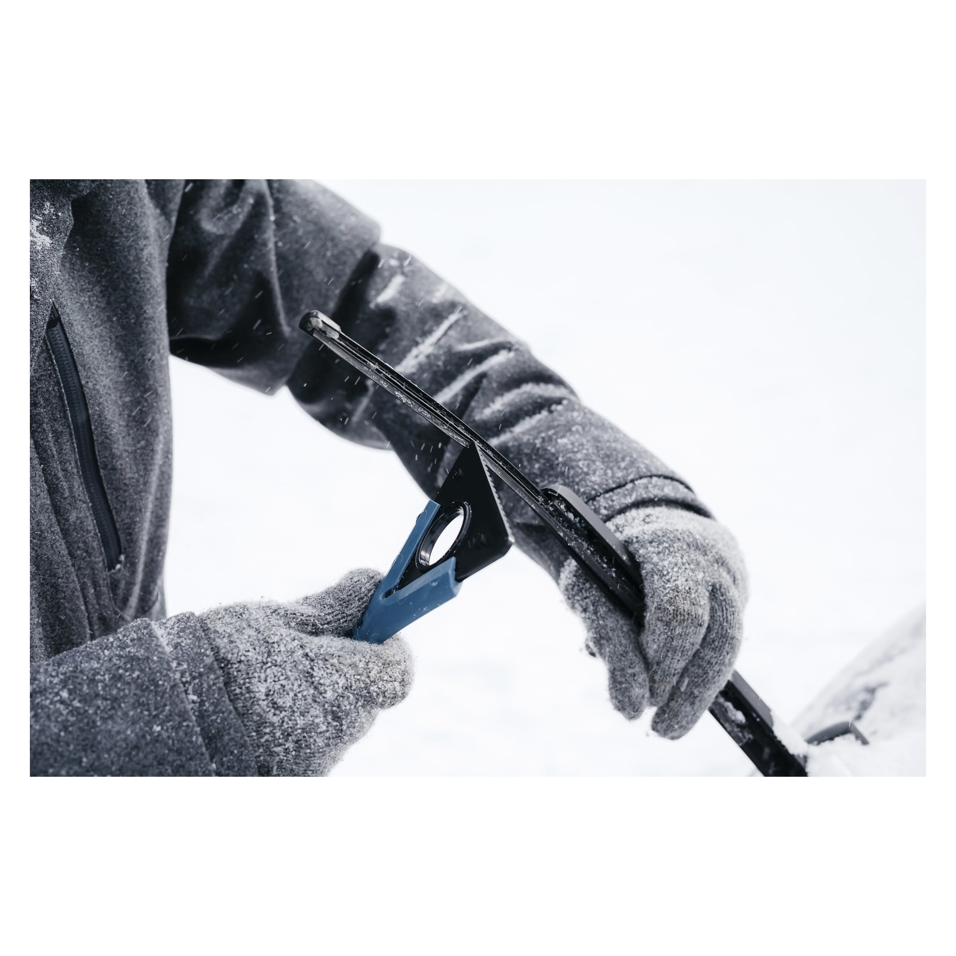 Någon i grå handskar och jacka använder Kungs Snow-busken med isskrapa MAX-IS för att rensa is från bilens vindrutetorkare i snötäckta, ljusa omgivningar. Scenen är täckt av snö.