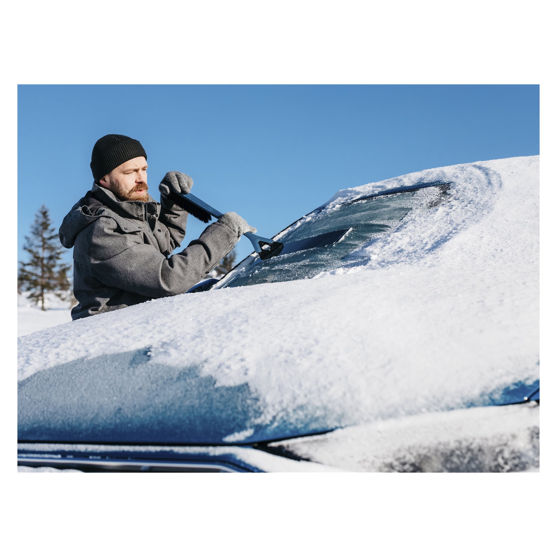 En man klädd i svart mössa, handskar och vinterjacka rensar snö och is från en bilruta med Kungs Snow-busken som visar en isskrapa MAX-IS på en solig, molnfri dag.