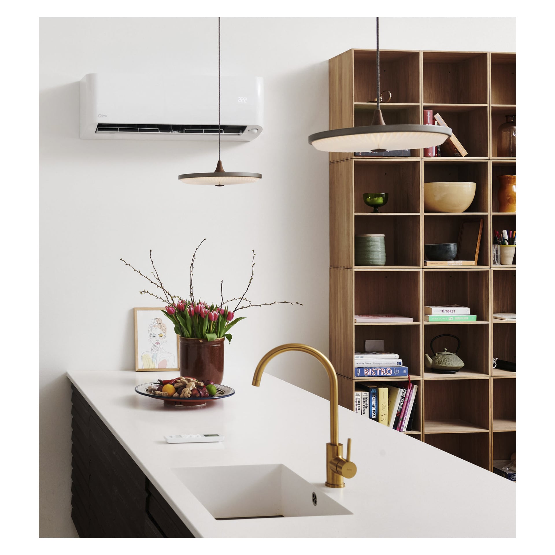 Modern kök med vit bänkskiva, mässingskran för diskhon, en vas med rosa tulpaner, två hänglampor ovanför och trähyllor med olika böcker, skålar och dekorativa föremål. Qlima Airs värmepump S-JA 7222 är installerad på väggen för klimatanläggning.