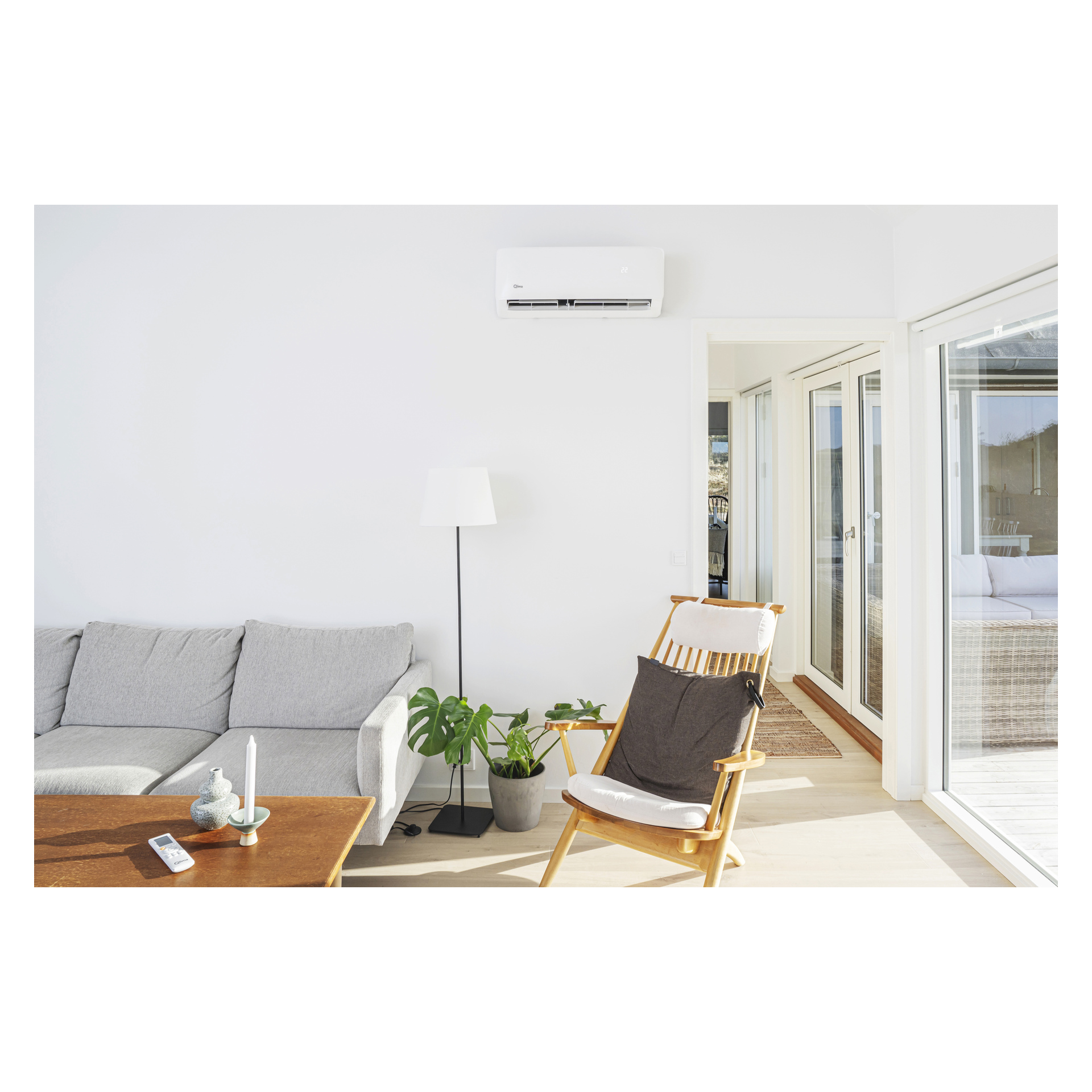 Ett stilfullt vardagsrum har en grå soffa, en trästol, en golvlampa och en krukväxt. En Qlima Air värmepump S-JA 2522 är installerad på väggen ovanför. Solljuset strömmar in genom stora glasdörrar, medan en fjärrkontroll vilar på det träbelagda soffbordet.