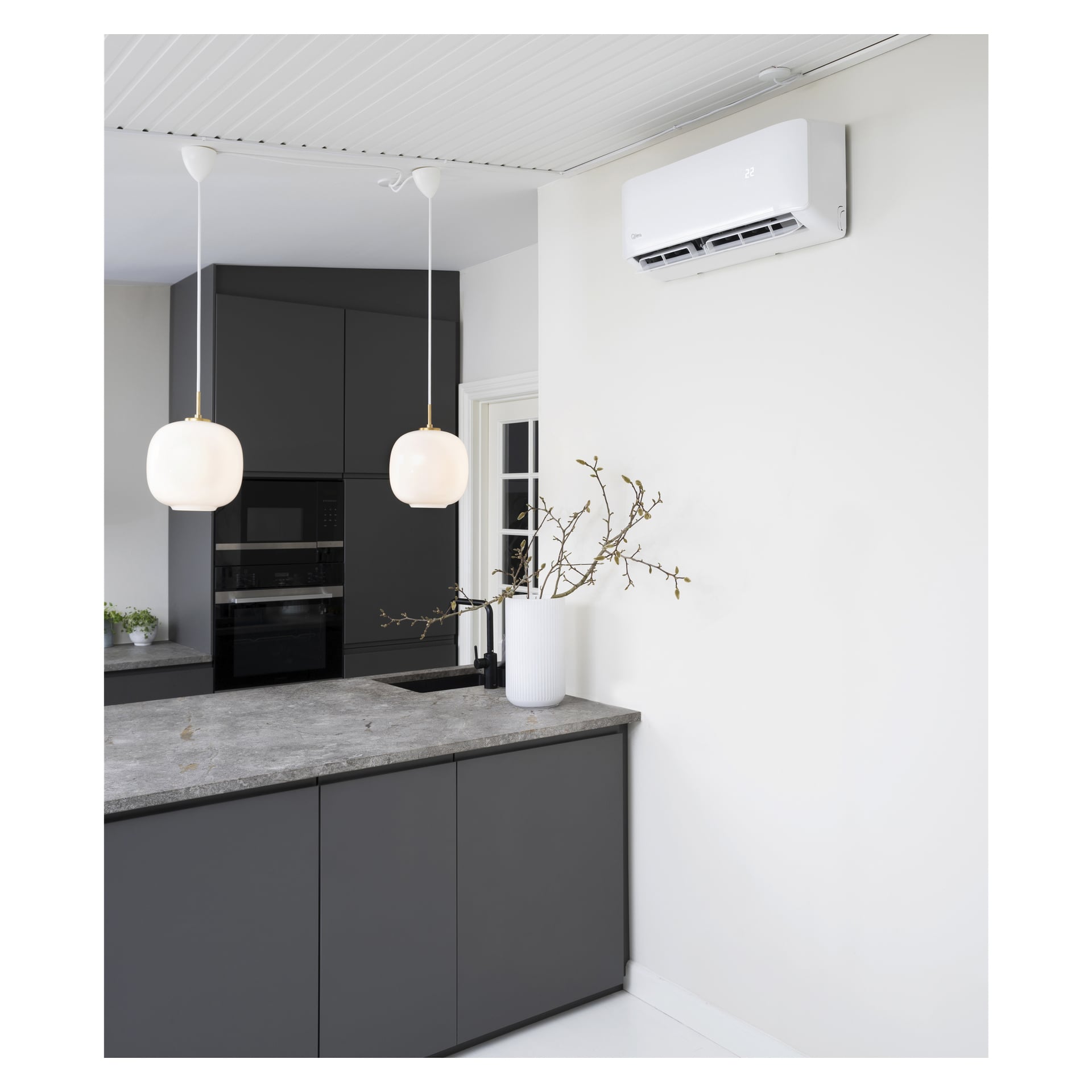 Samtida kök med mörka skåp, en grå bänkskiva och två hänglampor ovanför. En vas med grenar står på disken, medan Qlima Air Heat Pump S-JA 2522 är installerad ovanför. Den minimalistiska inredningen inkluderar vita väggar och en liten växt synlig i bakgrunden.