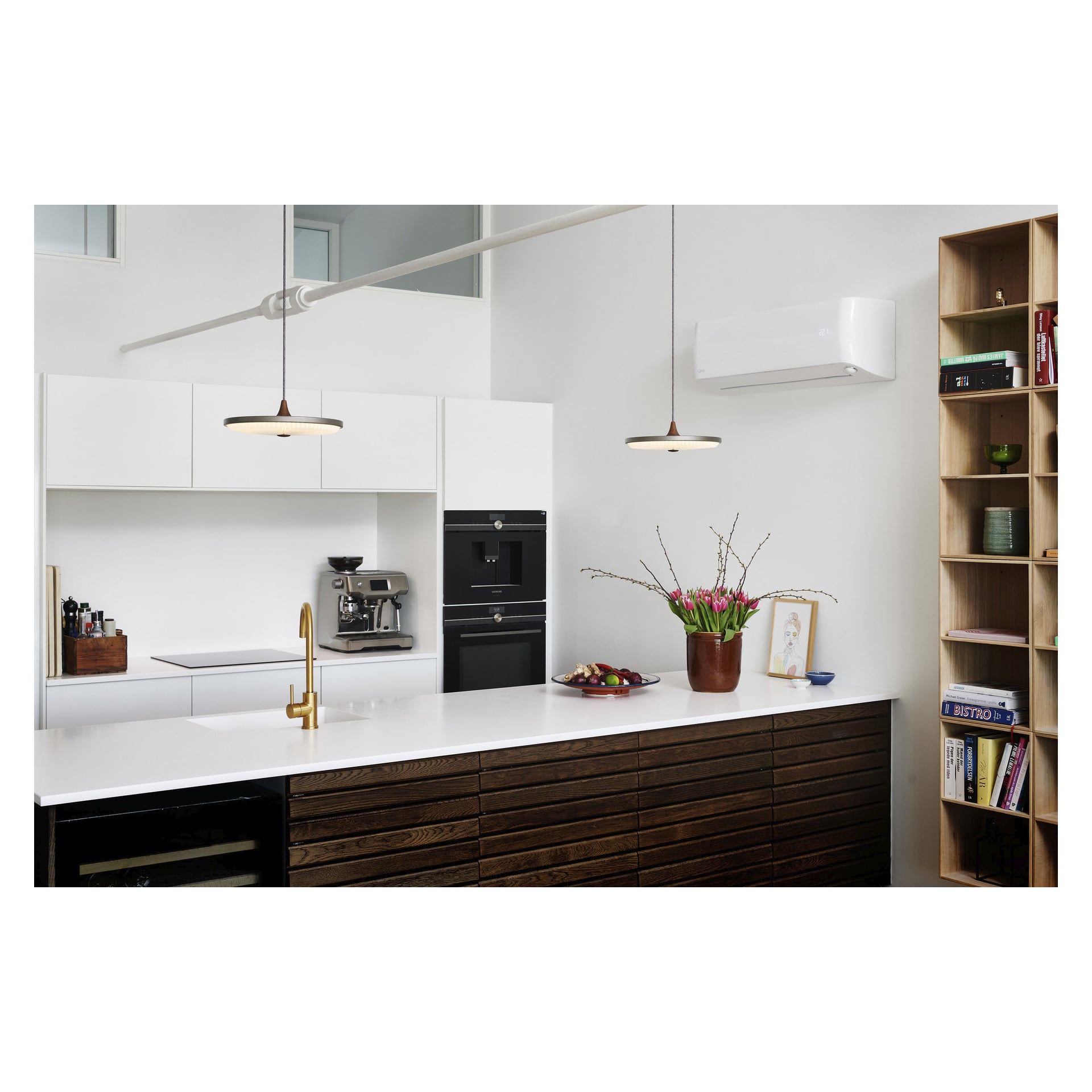 Det moderna köket med vita skåp, en mörk träö, en guldfärgad kran, inbyggd ugn, kaffemaskin, väggmonterade bokhyllor, två hänglampor ovanför, en fruktskål, färska blommor i en vas, inramad konst på bänkskivan och en luftvärmepump S-JA 7222 från Qlima.