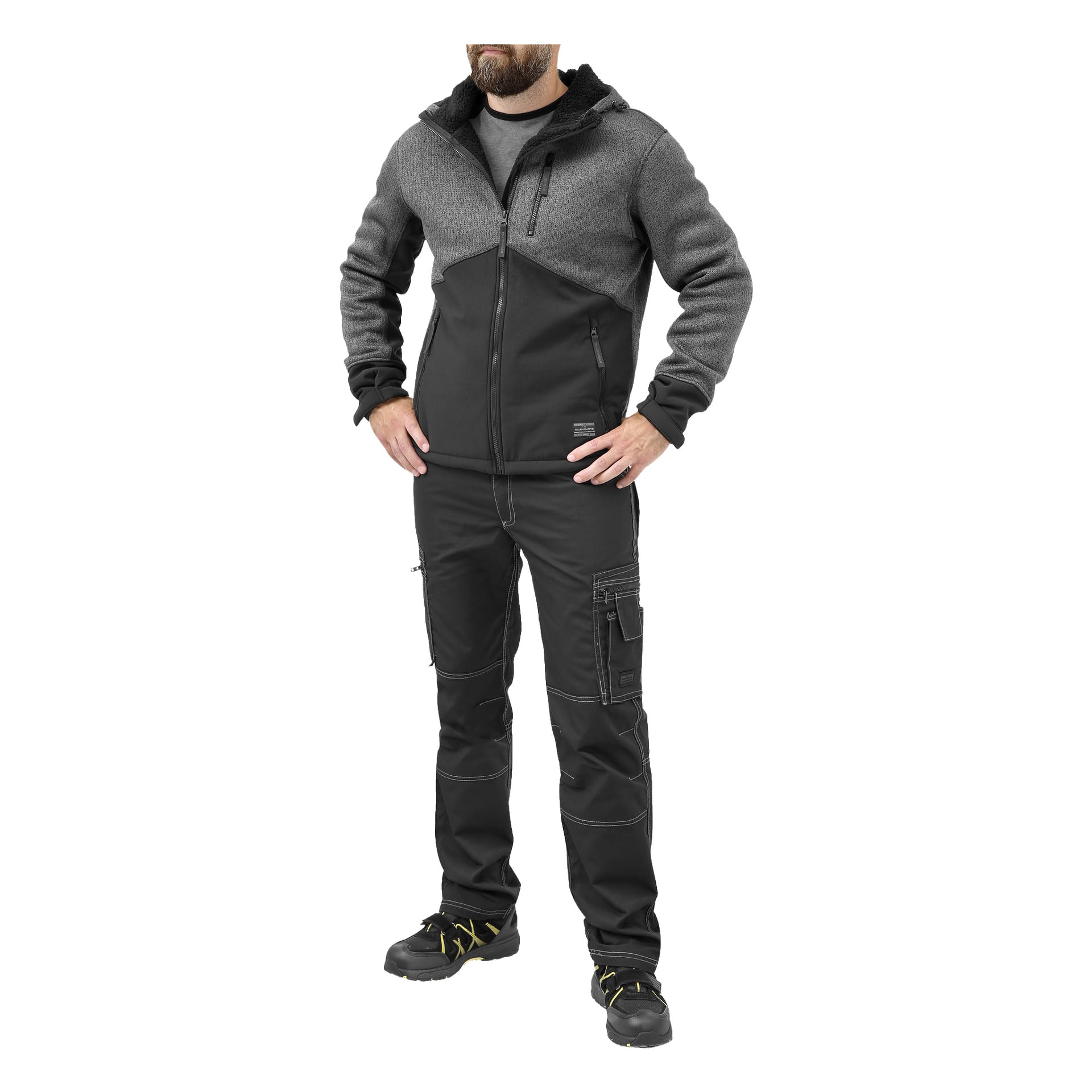 Mies seisoo kädet lanteilla, pukeutuneena harmaaseen ja mustaan Bluewear-miesten fleecetakkiin, mustiin cargo-housuihin taskuineen ja mustiin työkenkiin, joissa on keltaiset nauhat. Hänen päänsä on osittain pois kehyksestä.
