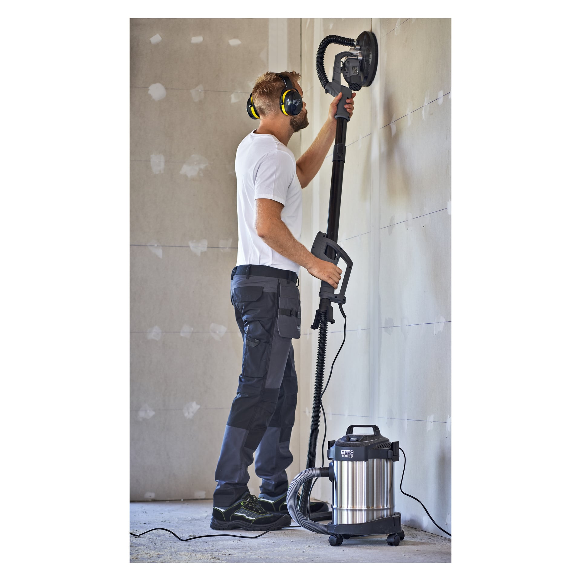 En mann i arbeidsklær og hørselvern pusser en uferdig vegg med en Meec Tools gipssliper WS-W HOME 300 koblet til en støvsuger.
