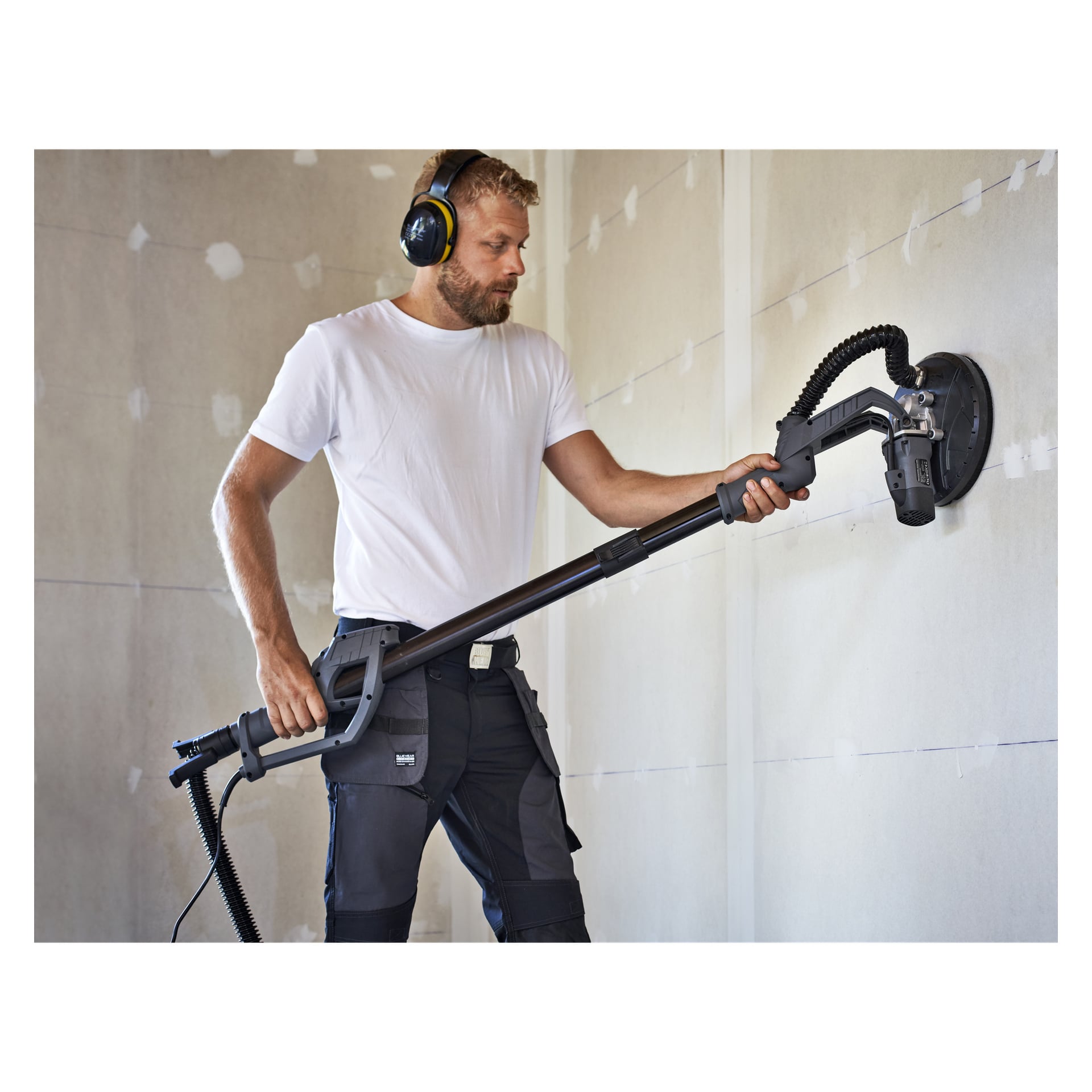 Iført hørselvern og en hvit t-skjorte pusser en mann en vegg i et byggeområde ved hjelp av Meec Tools Gipssliper WS-W HOME 300.