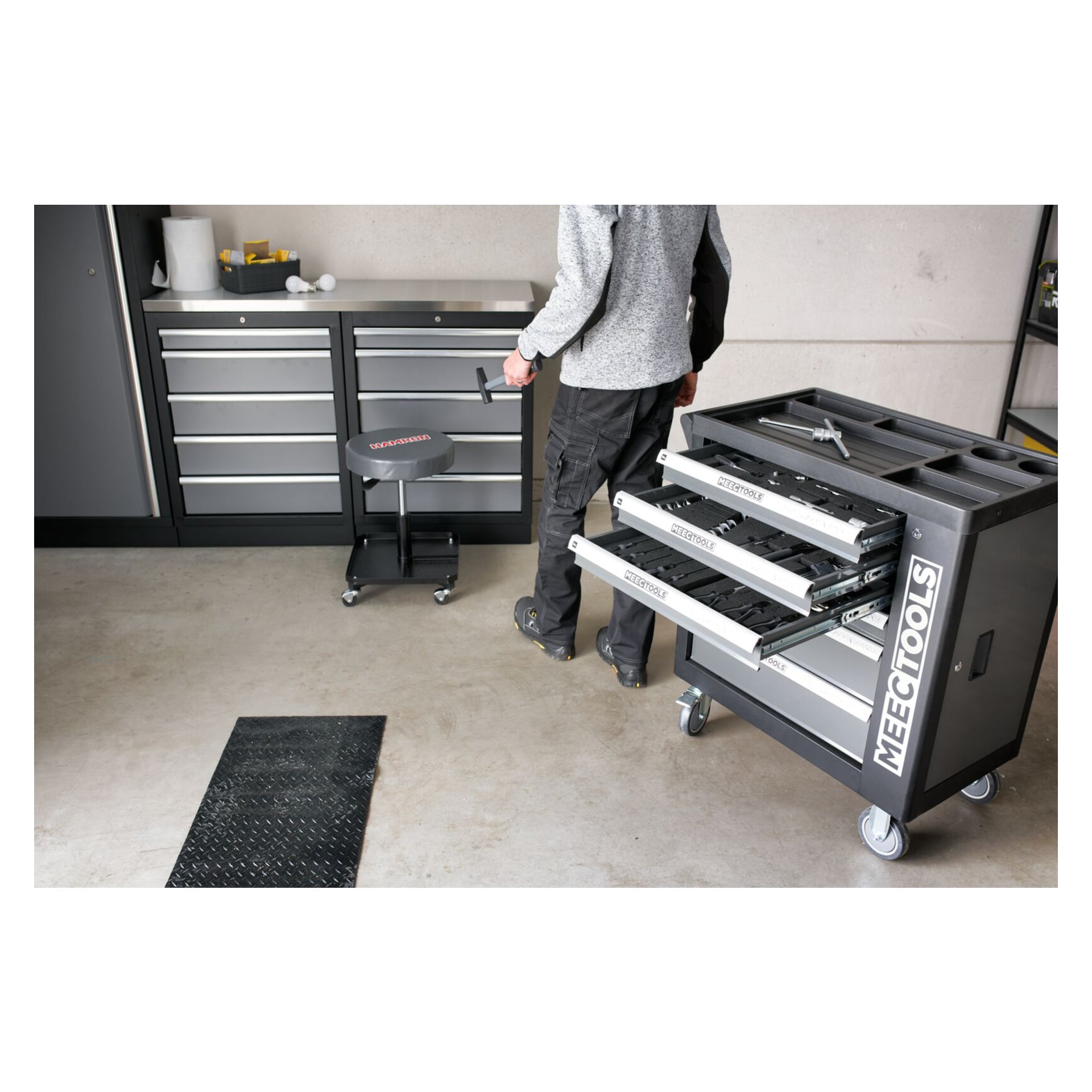 En person står bredvid en Meec Tools Tool Trolley med 167 delar, med tre lådor öppnade i en prydlig verkstad. I bakgrunden finns ett annat förvaringsskåp och en rullpall.