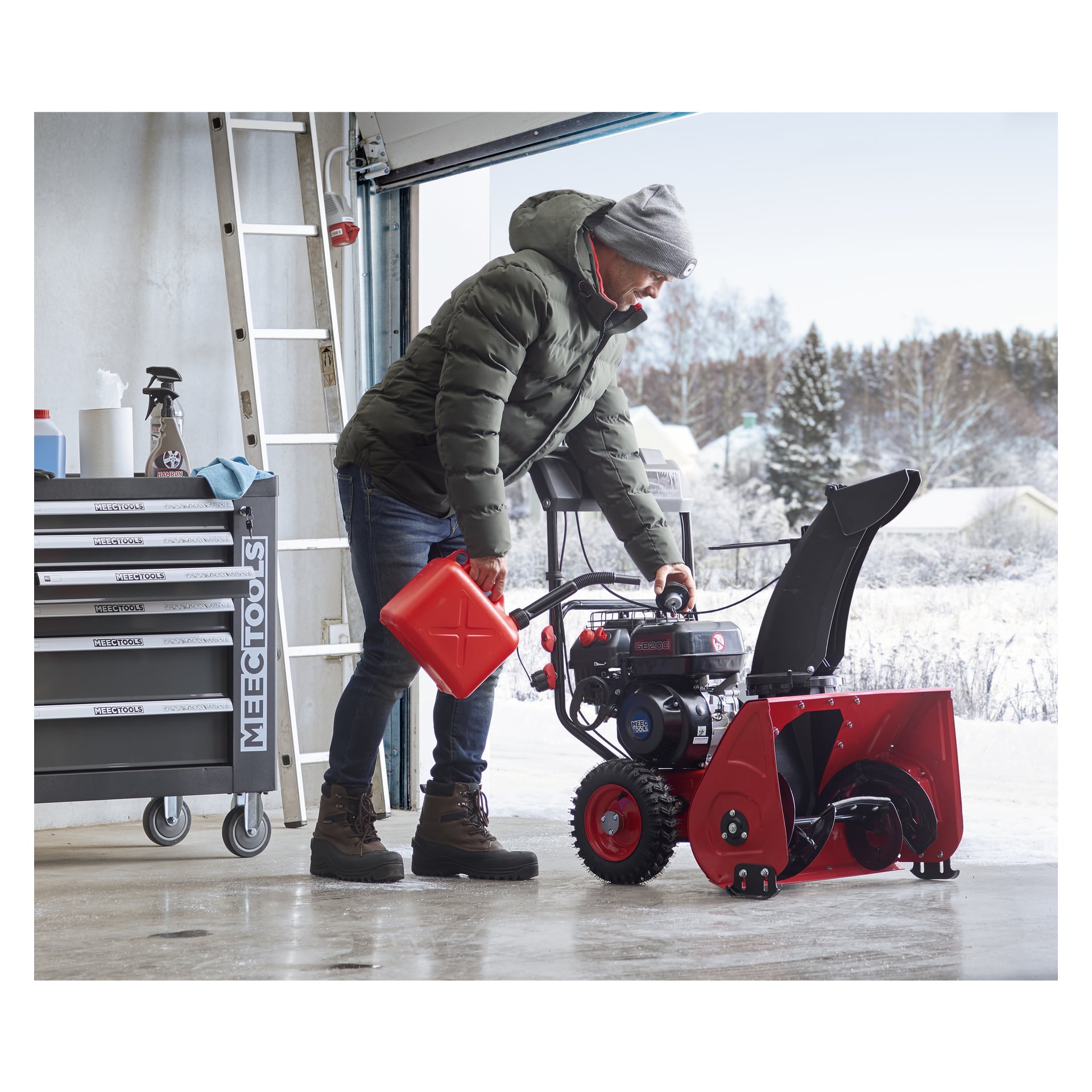 Klädd för vintern tankar en person en röd Meec Tools snöslunga med en röd bensindunk inne i ett garage. En verktygslåda står till vänster, medan man utanför det öppna garaget kan se snöiga träd och hus.
