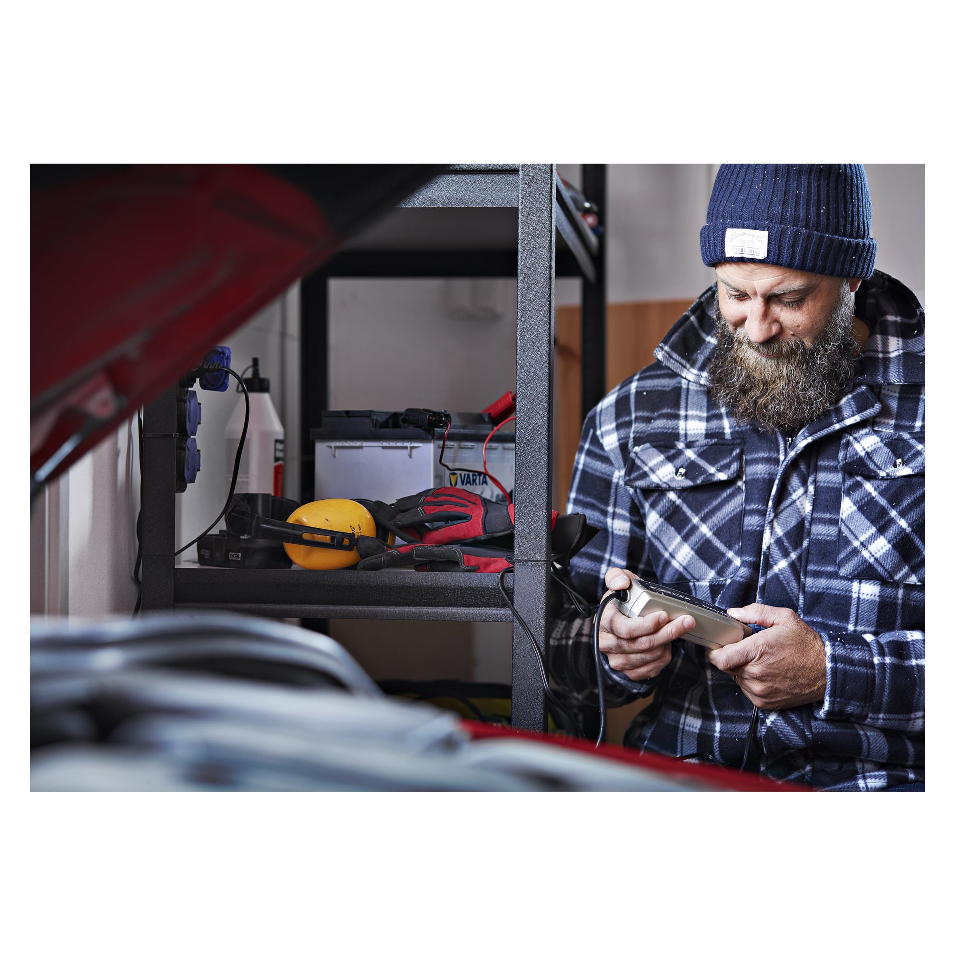 En man i rutig jacka och stickad mössa står i ett garage och håller i en CTEK batteriladdare MXS 3.8. Han står nära hyllor fyllda med verktyg och utrustning, medan huven på en röd bil är öppen framför honom.