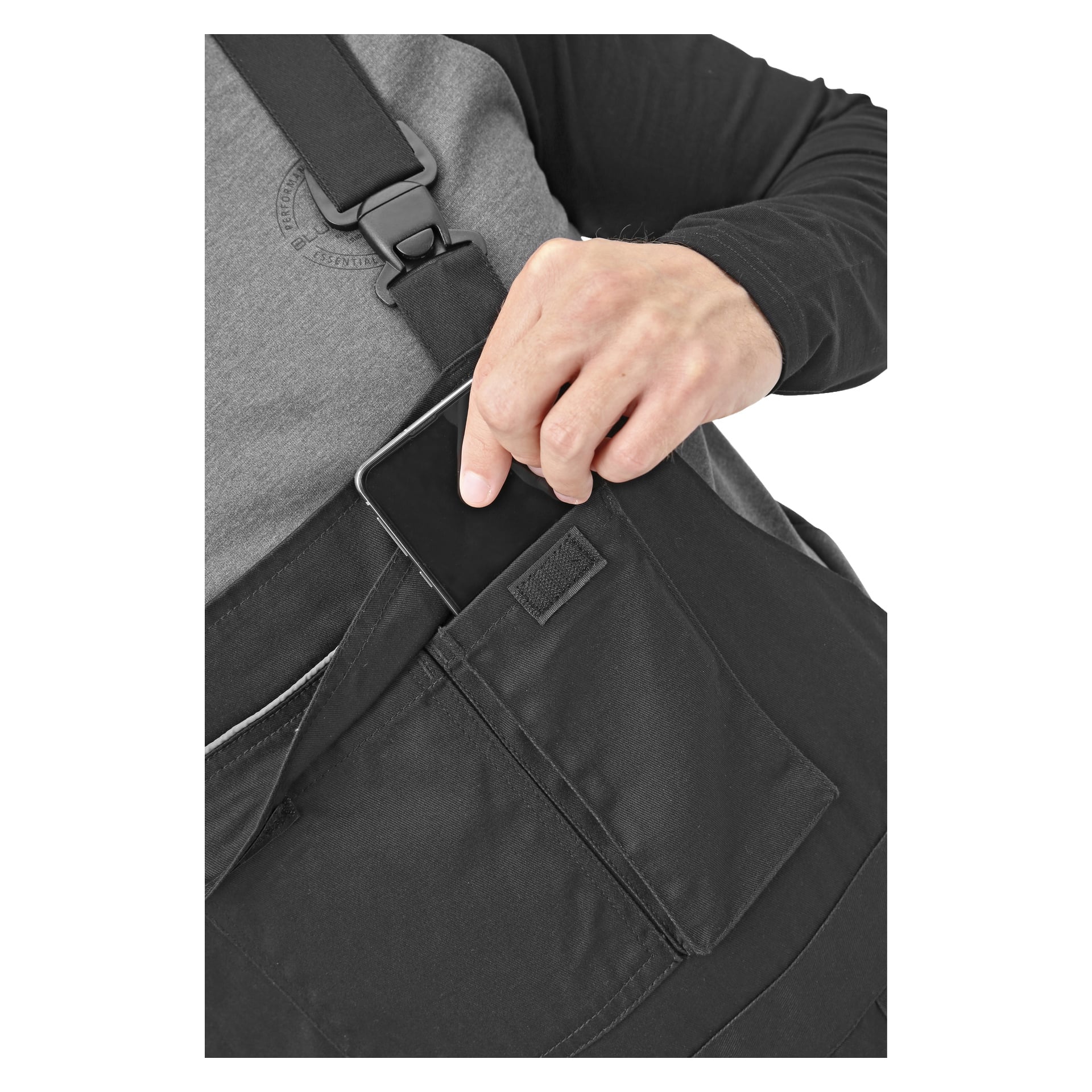 En person stoppar en svart smartphone i framfickan på Bluewear herrarnas Harry-overall. Endast deras överkropp och hand syns, och de är klädda i en långärmad skjorta i svart och grå färger.
