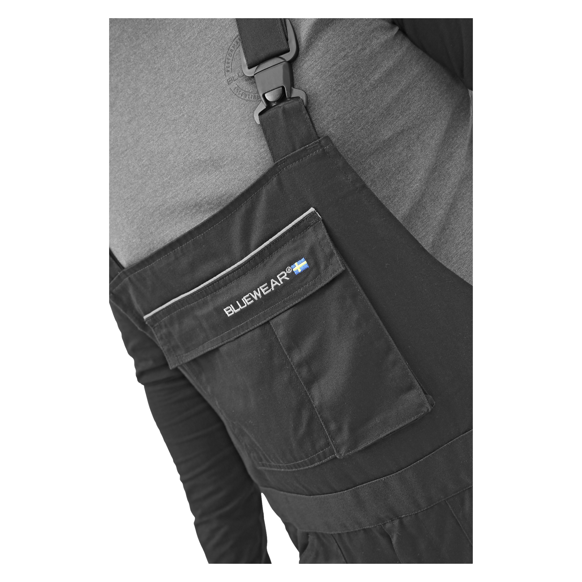 En närbild visar en person klädd i Bluewears Harry-herroveraller, som har en framficka med en flik som visar Bluewear-loggan och en liten svensk flagga. Personen har på sig en grå skjorta under hängselbyxorna.
