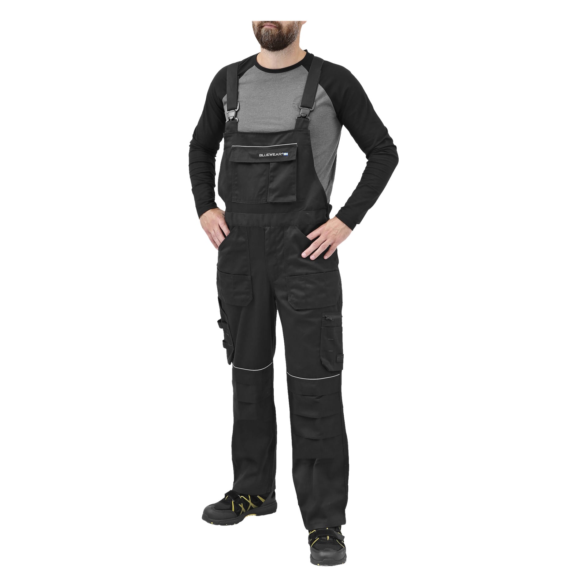 En skäggig person står med händerna i sidan, klädd i Bluewear Men's Harry-overaller – svarta arbetsplagg med flera fickor – tillsammans med en grå och svart långärmad skjorta och svarta arbetsskor med gula accenter. Skägget döljer delvis ansiktet.
