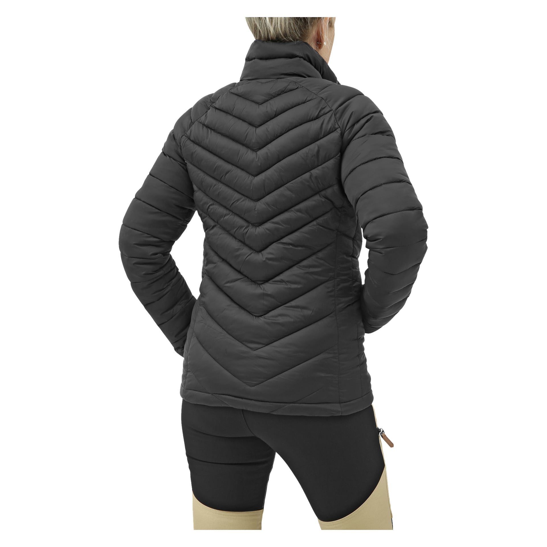 En person vänder blicken bort från kameran med händerna på höfterna, klädd i en svart Kayoba Outdoor Women's lättviktsjacka med ett vadderad chevron-design på ryggen, och bär svarta och beige byxor.