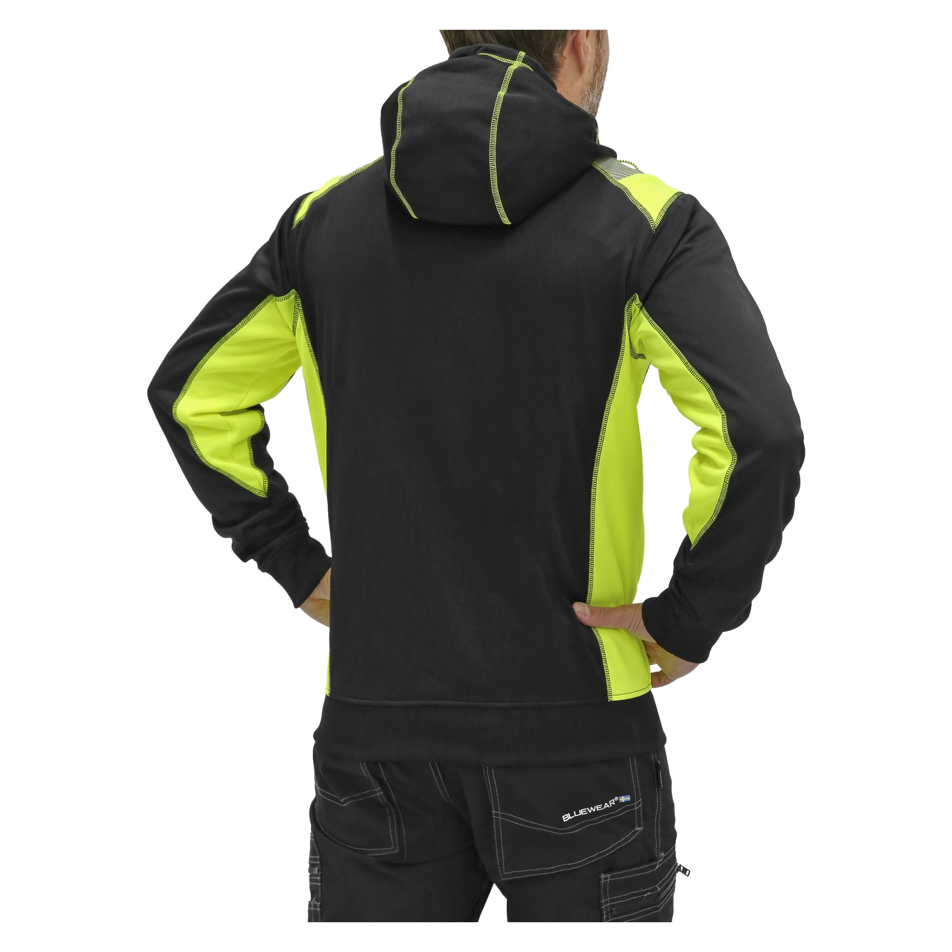 En person står med ryggen mot kameran, händerna vilande på höfterna, klädd i en Bluewear Men's hoodie Felix i svart och neongult, tillsammans med svarta byxor framhävda med vita sömmar.