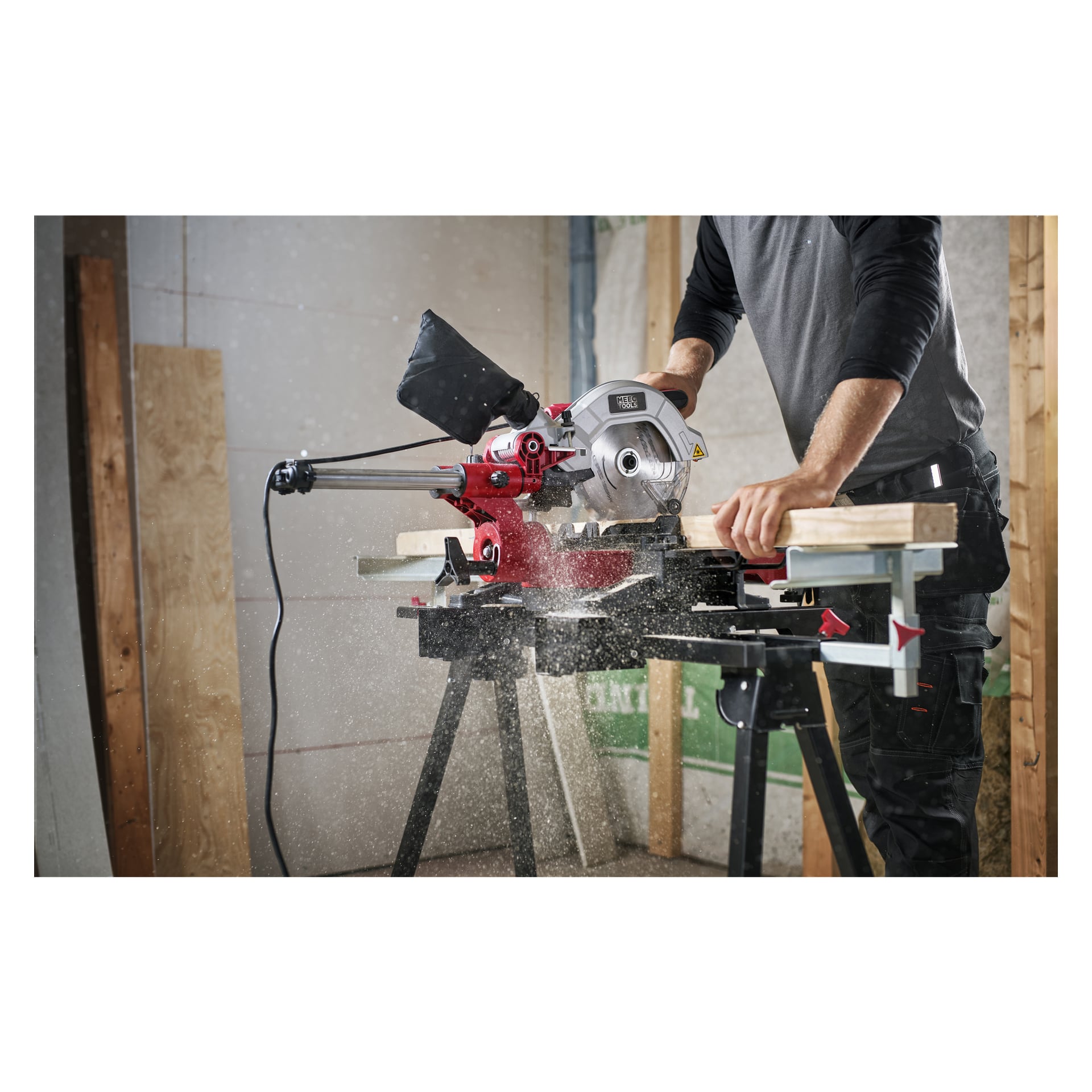 Henkilö käyttää Meec Tools Mitre -sahaa puun leikkaamiseen työpöydällä työpajassa, ja sahanpuru näkyy ilmassa. Henkilöllä on päällään pitkähihainen paita ja tummat housut.