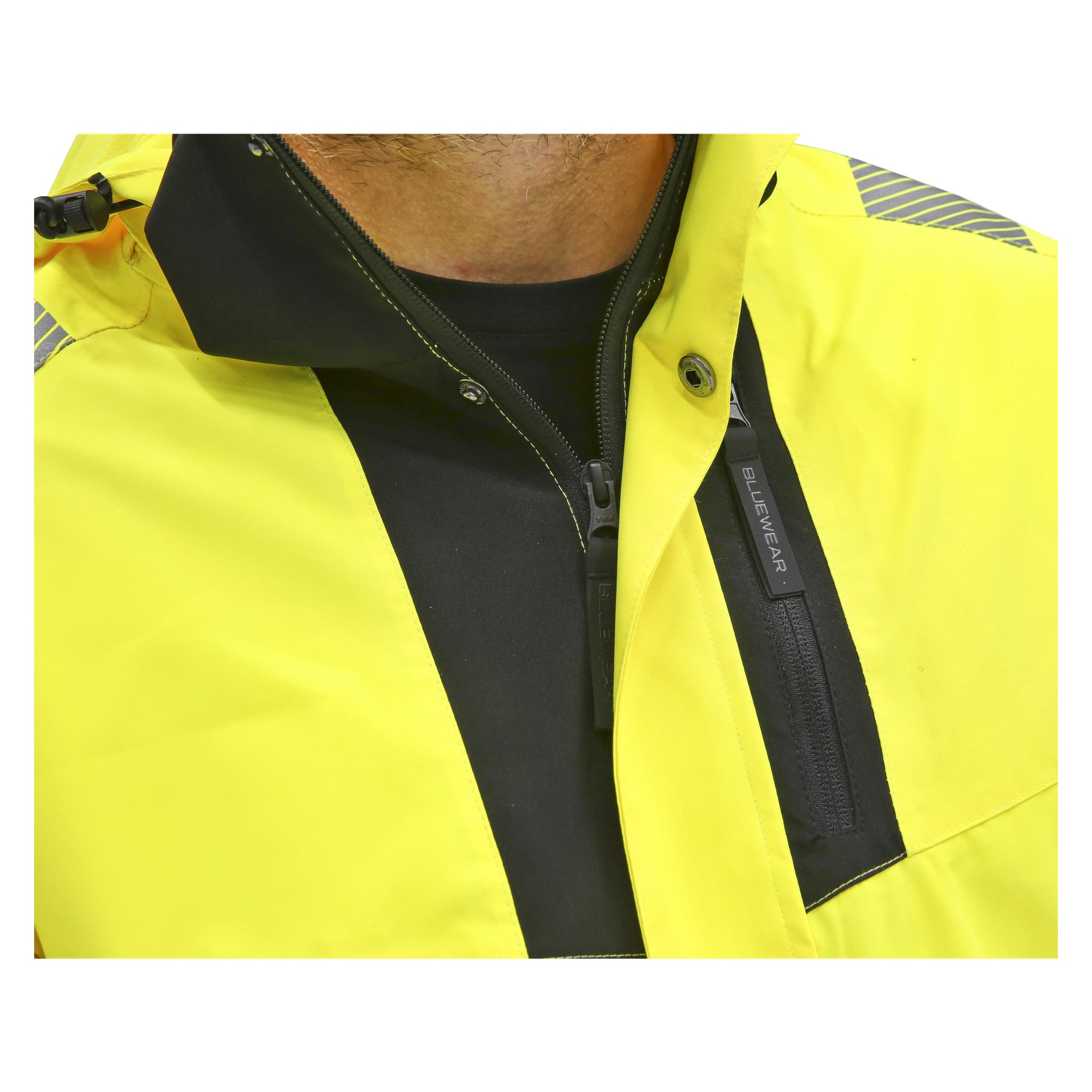 En närbild visar någon i en Bluewear Hi-Vis-jacka, kännetecknad av sin klargula färg med svarta detaljer och en bröstficka med Bluewear-logotypen. Personen bär också en svart skjorta under jackan, där endast halsen och övre bröstet syns.