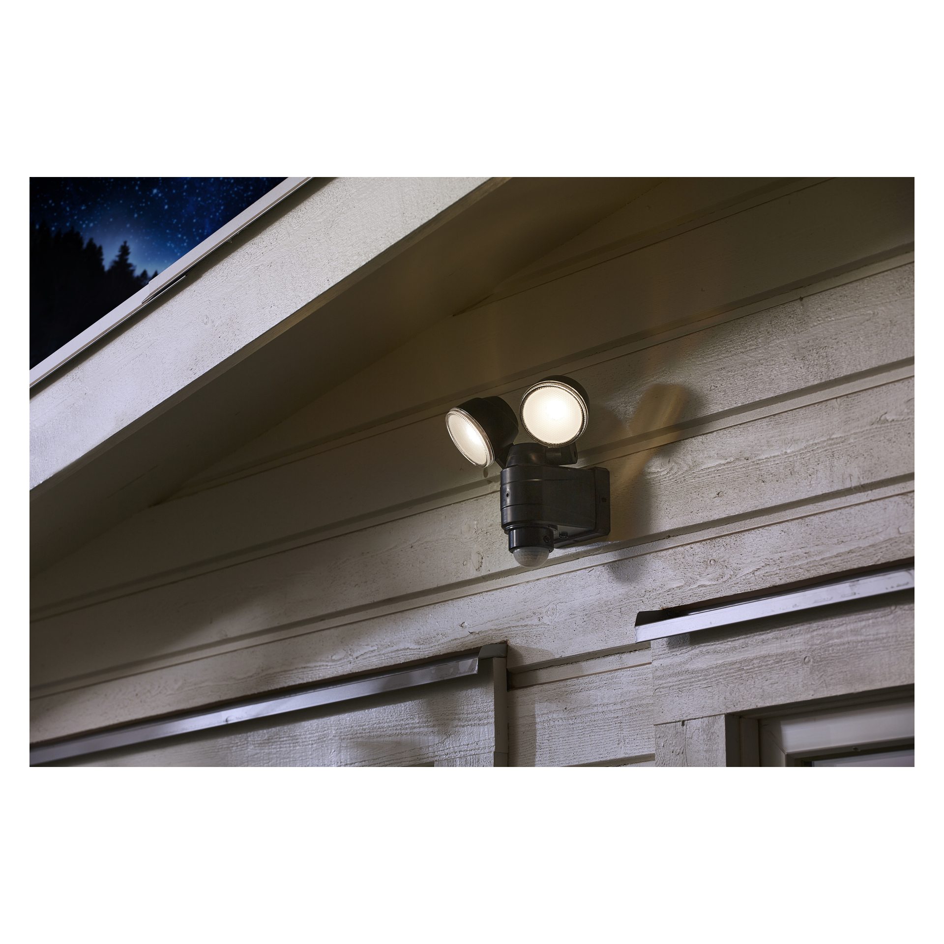 En Anslut-strålkastare med LED-rörelsedetektor är installerad under takfoten på husets yttervägg och lyser starkt på natten med sina två runda lampor, medan en del av den mörka himlen syns i ett hörn.