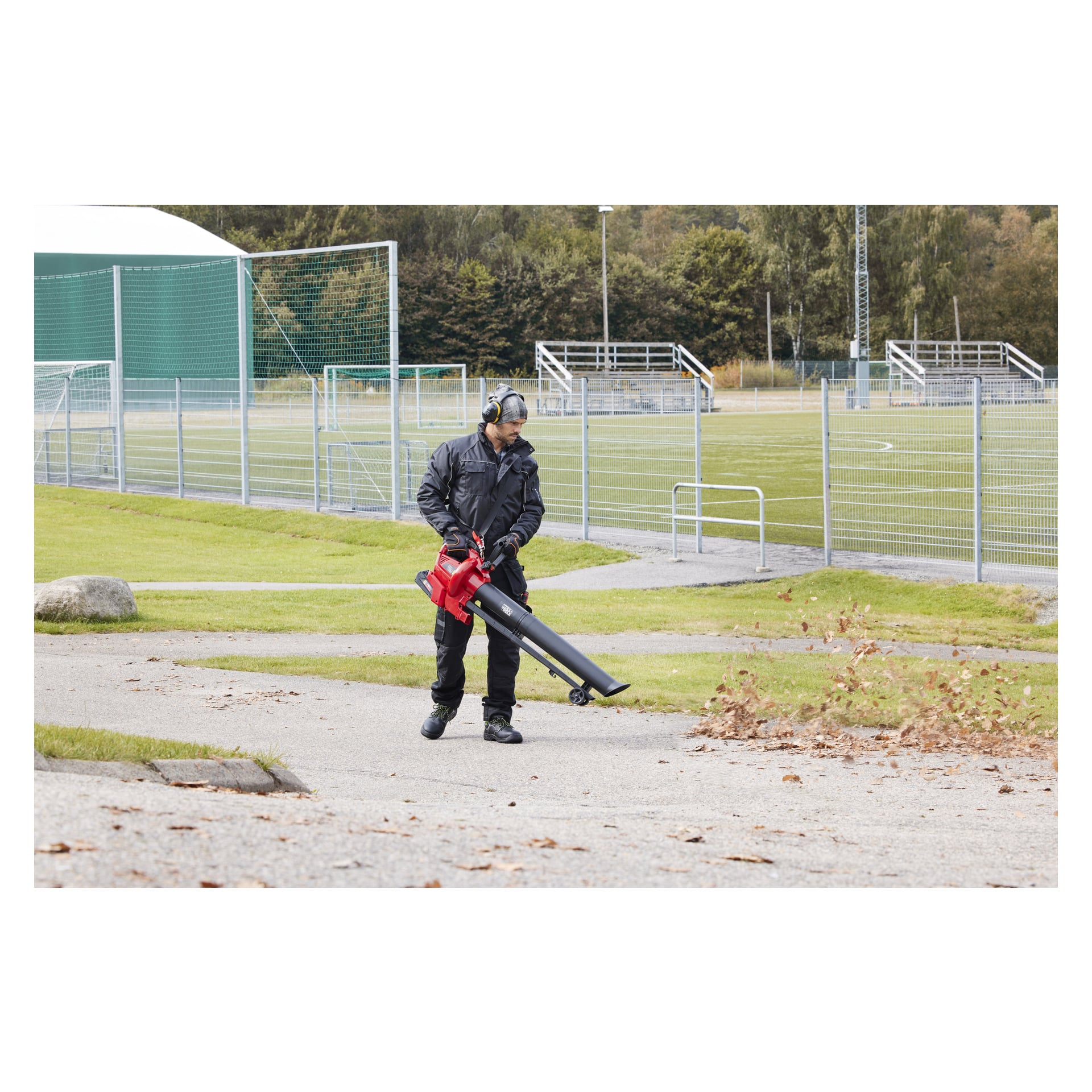 En person i skyddsutrustning använder en Meec Tools Multiseries 40 V bladblås/sug, där löv tas bort från en asfalterad yta bredvid en sportplan omgiven av tomma läktare och grönt staket.