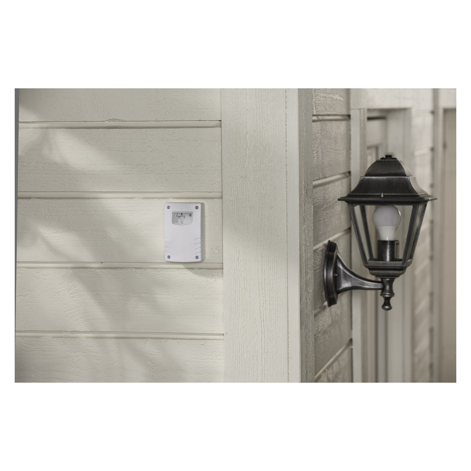 Ett Anslut Twilight-relä är installerat på beige panel bredvid en svart utomhuslampa med en vit glödlampa och en vit plastanordning som kan vara en termostat eller säkerhetssensor.
