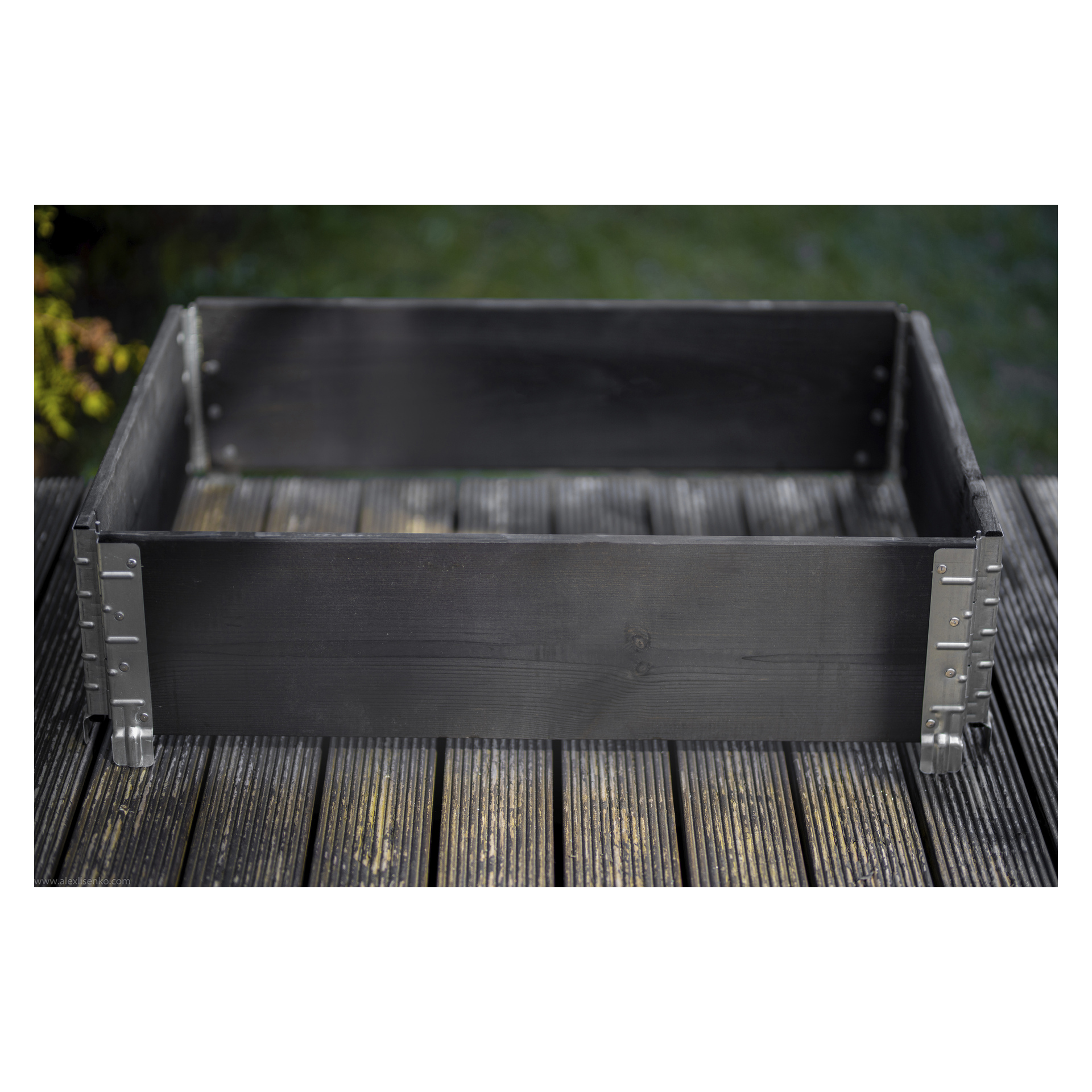 Et svart rektangulært Hard Head Cultivation Collar med metallbraketter i hjørnene hviler tomt på en treplate med lameller utenfor, omgitt av gress og annet grønt.