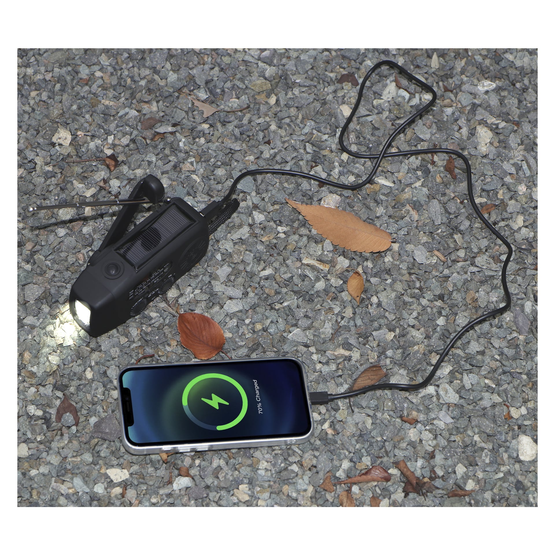 En livfull uppdragbar FM-radio med solpanel laddar en smartphone via en USB-kabel. Ficklampan lyser upp grus och några torkade löv i närheten. Smartphonens display visar att den laddar.
