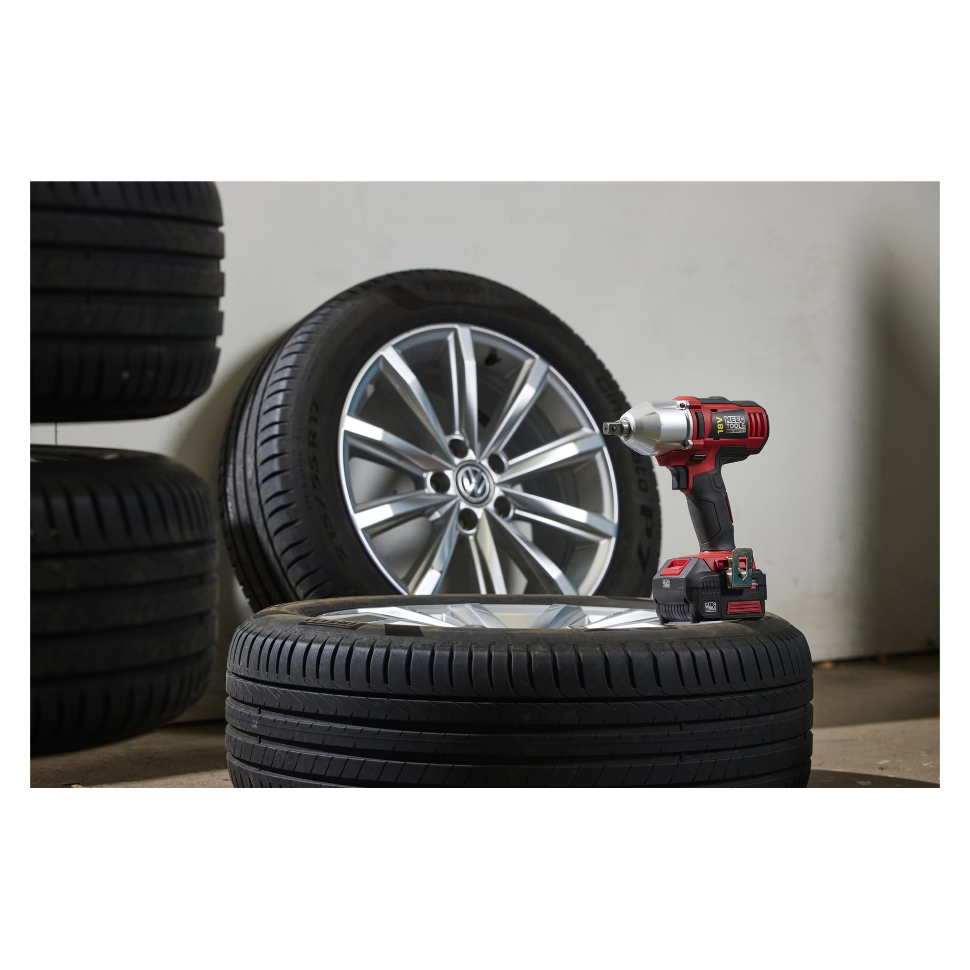 Punamusta Meec Tools Multiseries IW-18V EASY 100 -akkukäyttöinen iskuavain istuu hopeisen kevytmetallivanteen renkaan päällä. Lisää renkaita on pinottu lähelle, taustalla näkyy seinä.